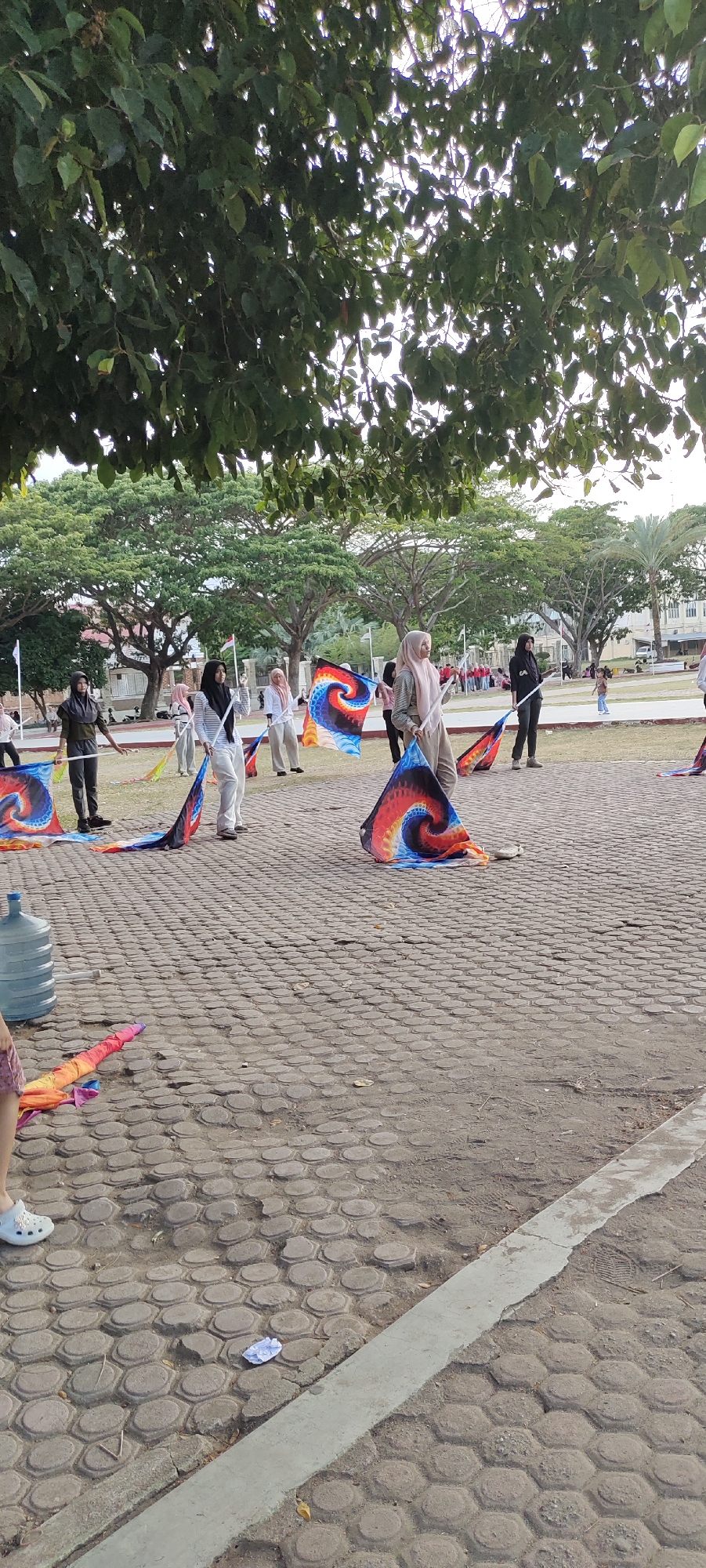 The image depicts an outdoor scene, likely in Lhokseumawe, Indonesia, featuring a group of individuals practicing flag routines. The ground is paved with interlocking hexagonal stones. Several people, predominantly women wearing headscarves, are holding and manipulating large, colorful flags with a spiral design. The flags are vibrant, with swirling patterns of red, blue, yellow, and orange. Some flags are raised, while others are spread on the ground. In the background, lush green trees provide a canopy, and more distant foliage and buildings are visible, suggesting an urban or park-like setting. A large blue water cooler is positioned on the left side of the frame, near a colorful, folded fabric on the ground. The lighting suggests it is daytime, possibly late afternoon, given the soft, diffused light. The overall atmosphere appears to be one of organized activity or practice.