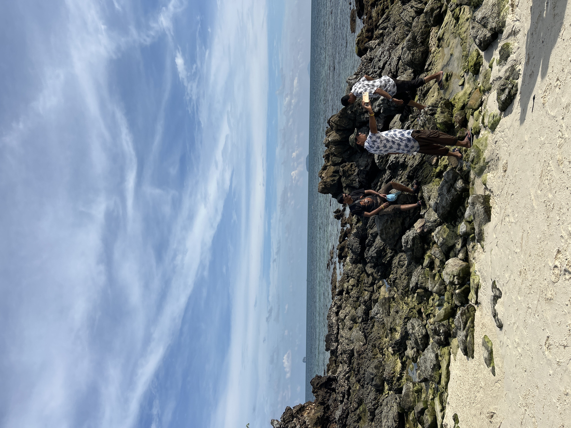 The image shows a rocky shoreline with white sand in Tambon Phru Nai, Thailand. Three individuals are present on the rocks. One person is seated on the left, wearing a black t-shirt and shorts, holding a light blue mask. In the center, another person, wearing a patterned shirt, a camouflage cap, and brown pants, is holding up a smartphone to take a picture, with their arm extended towards the right. To the right, a third person, dressed in a patterned shirt and black shorts, is facing away from the camera, seemingly walking towards the sea. The background features the ocean under a blue sky with wispy white clouds, and distant islands can be seen on the horizon. The rocks are dark and rugged, with patches of green algae or moss.