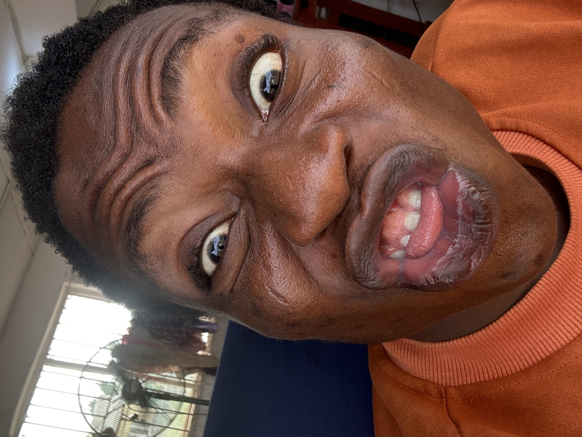 A close-up, angled shot depicts a person with dark skin, curly hair, and an orange sweater making a distorted facial expression. The eyes are wide and the mouth is open, revealing teeth and tongue. Forehead wrinkles are prominent. The background includes a window with blinds, a fan, and a blue surface. The location is unknown.