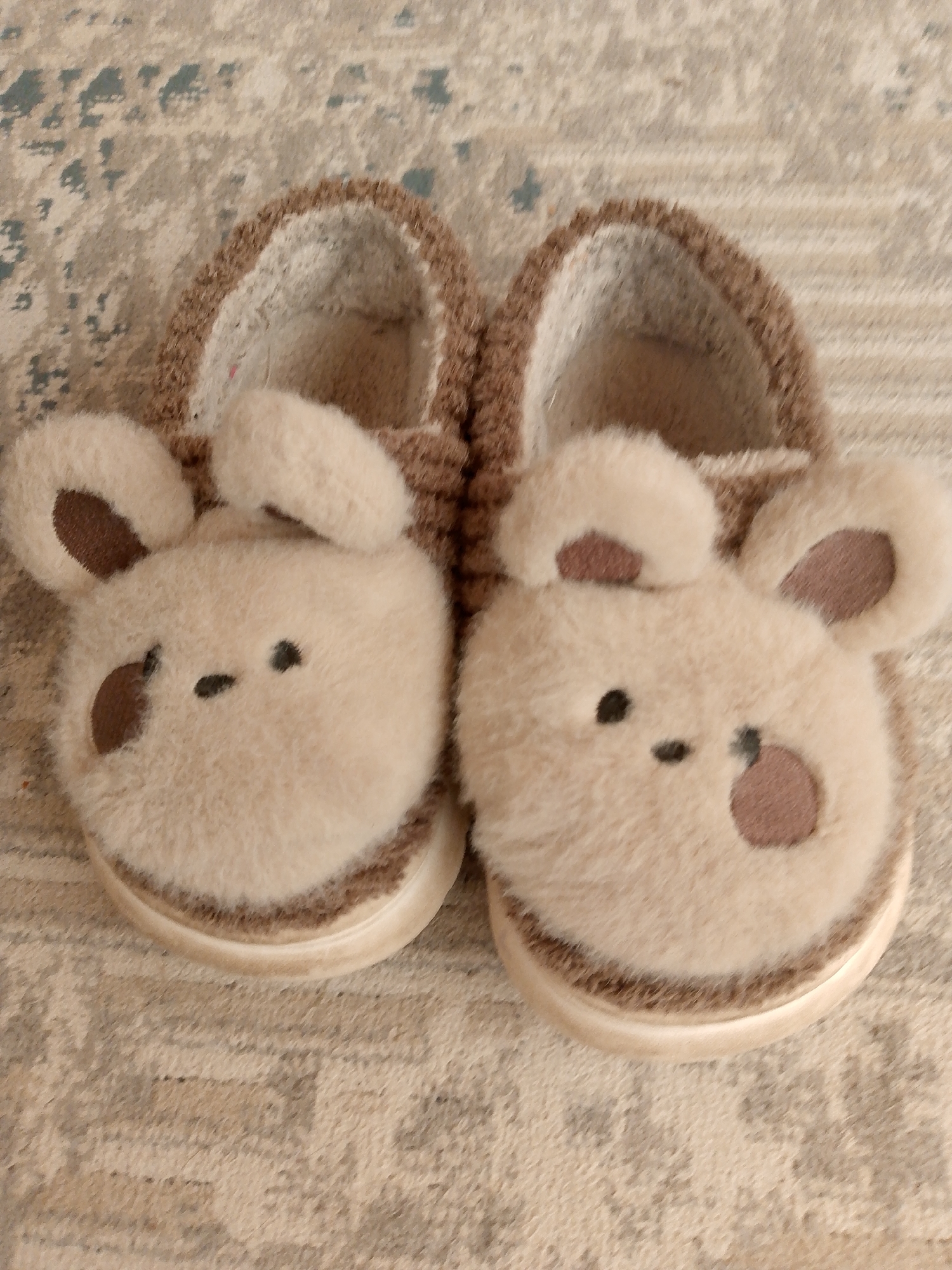 The image shows a pair of fluffy, beige, bear-shaped slippers resting on a textured rug. The slippers have round ears with brown accents and embroidered facial features consisting of small black dots for eyes and a dash for a mouth. One slipper also has a brown accent on its cheek. The rug appears to be a light brown or beige color with a subtle pattern of darker lines and shapes. The slippers are positioned side-by-side, with the left slipper slightly overlapping the right. The overall impression is one of comfort and cuteness. There is no indication of the city of Prigorognoye, Kyrgyzstan in the image.