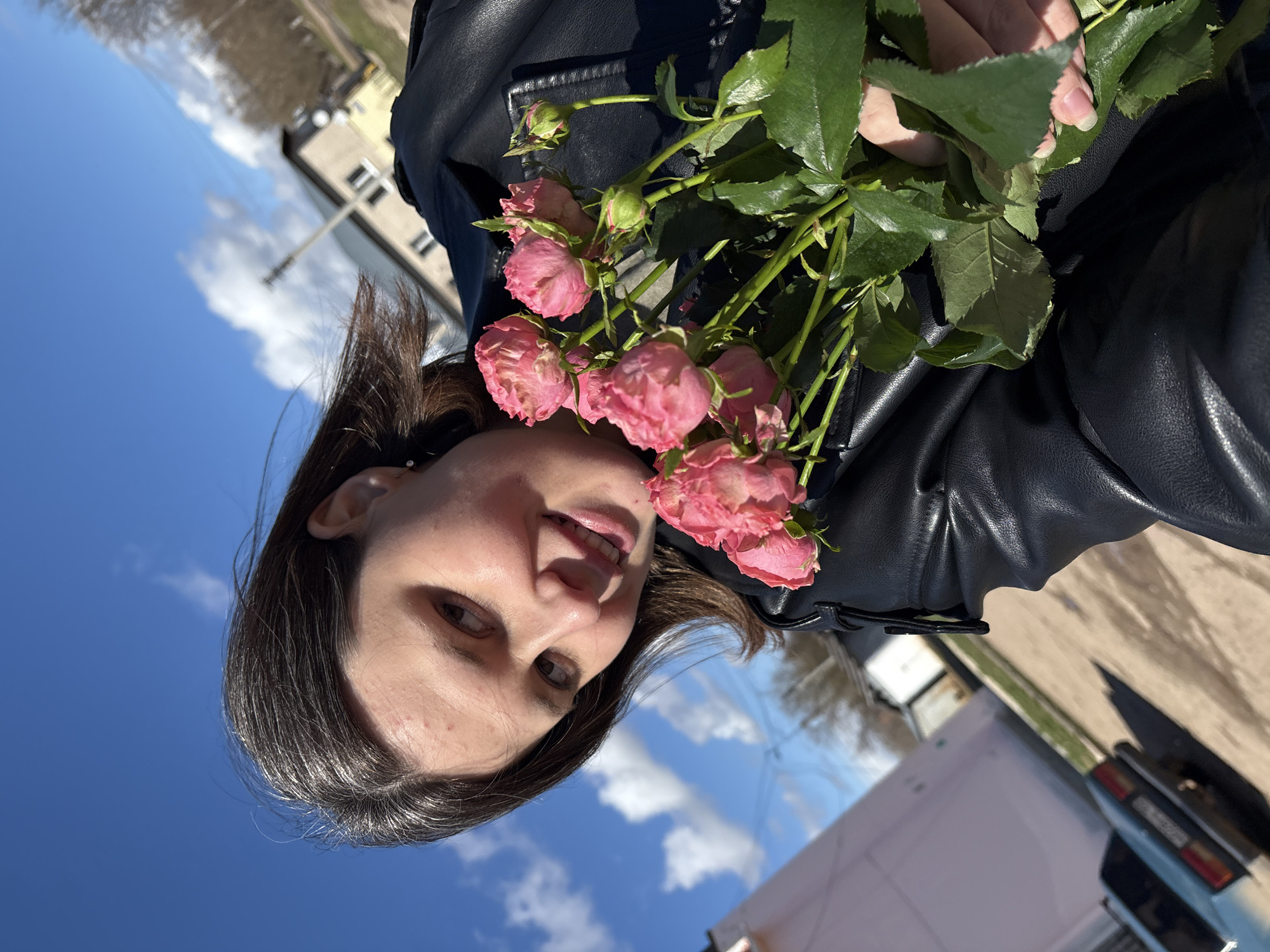 The image, appearing rotated 90 degrees counter-clockwise, shows a young woman outdoors holding a bouquet of pink roses. She has dark hair, brown eyes, and a slight smile as she looks towards the camera. She is wearing a black leather jacket. The bouquet consists of several small, vibrant pink roses with green leaves, held close to her face and shoulder.
The background features a clear, bright blue sky with scattered white, fluffy clouds, indicating sunny weather during the daytime. In the mid-ground, there are buildings, possibly residential, and a paved area or road. Trees with bare branches are visible in the distance, suggesting early spring or late autumn. Parts of vehicles, including a white van or truck and the rear lights of another car, are visible on the right side of the frame. The woman appears cheerful. There is no visible text in the media file. The setting is an outdoor, possibly urban or suburban environment, in Фоки, Russia.