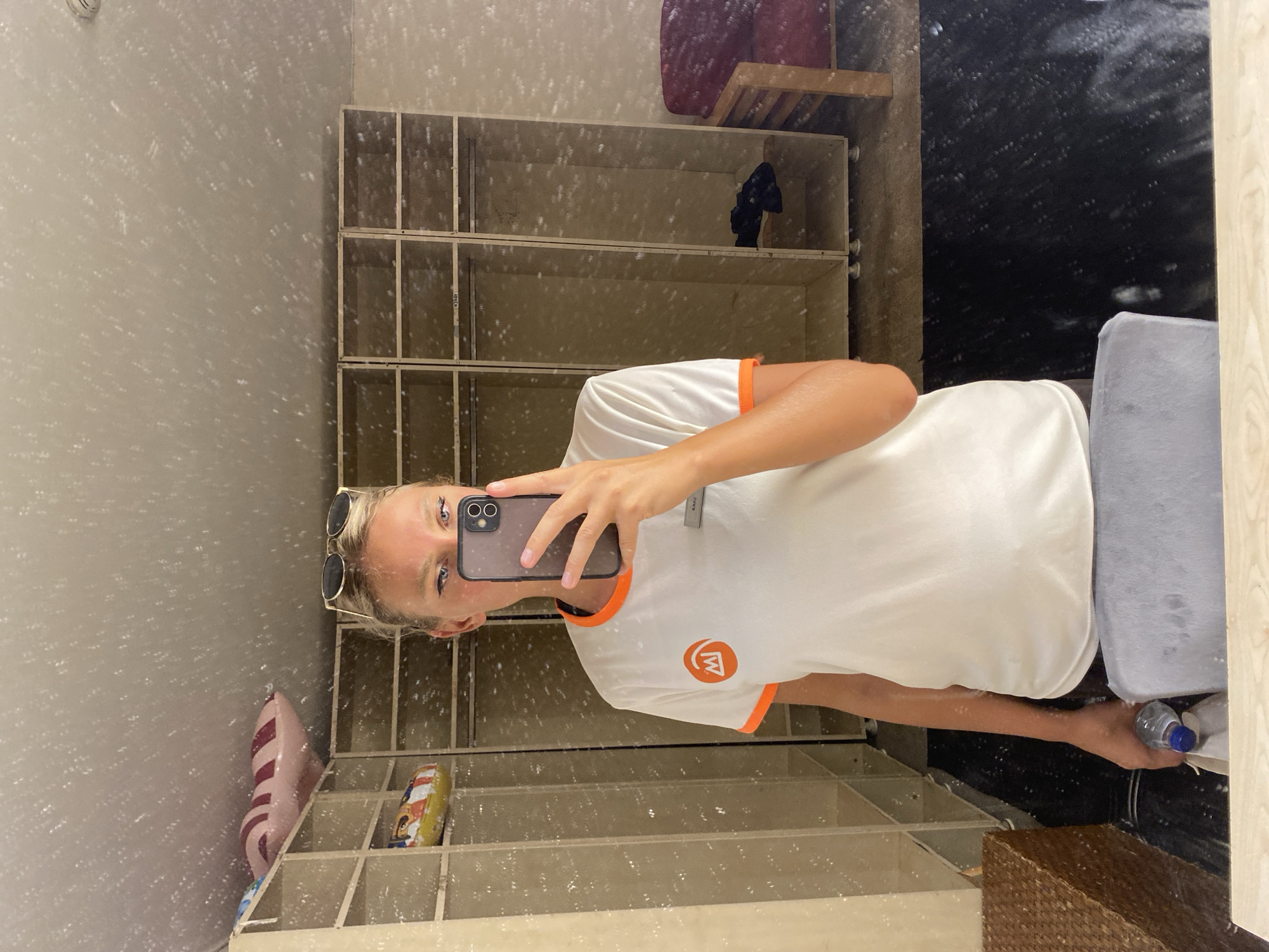 A young Caucasian woman with blonde hair pulled back and sunglasses perched on her head takes a mirror selfie in what appears to be a locker room. She is wearing a white t-shirt with orange trim and a circular orange logo on the left chest. Her right hand holds a smartphone, capturing the reflection. In her left hand, she holds a clear plastic water bottle. The background of the reflection shows a row of empty wooden lockers and a red upholstered chair. The environment appears somewhat cluttered with various items on shelves and the floor. The overall lighting is bright, and there are numerous light specks visible on the mirror's surface, possibly dust or imperfections. There is no explicit information to indicate this is in Aksu, Turkey.