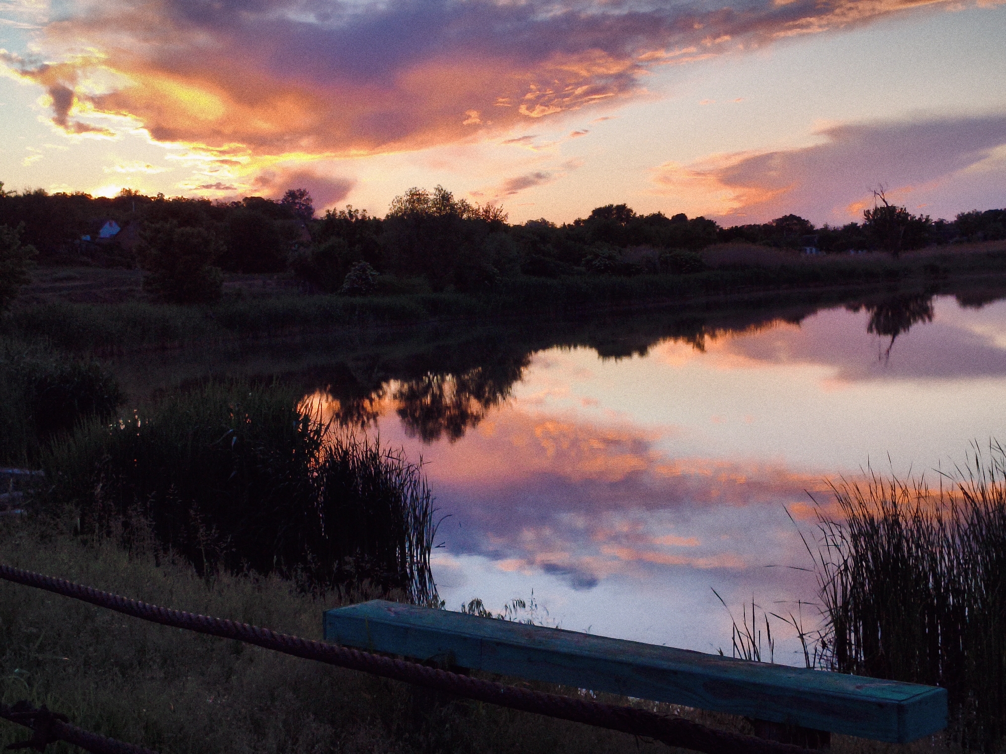 The image shows a serene sunset over a body of water, likely a lake or a wide section of a river, located in Ustyvytsia, Ukraine. The sky is filled with clouds, painted in hues of orange, pink, and purple by the setting sun. The trees and vegetation along the bank are silhouetted against the colorful sky. The water reflects the sky's colors, creating a mirror-like effect. In the foreground, there is a rustic barrier, consisting of a weathered wooden beam painted in a muted green color, and a rusty rope, that is partially obscured by vegetation. The overall scene evokes a sense of tranquility and natural beauty.