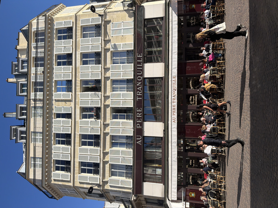 The image displays the multi-story facade of "Au Père Tranquille," a brasserie located at 16 Rue Pierre Lescot, 75001 Paris, France. The building is of classic Haussmannian architectural style, featuring a light-colored stone exterior, multiple floors with evenly spaced windows fitted with light grey shutters (some partially open), and wrought-iron balconies on upper levels. A mansard roof with dormer windows tops the structure. The ground and first floors are occupied by the brasserie, identifiable by "AU PERE TRANQUILLE" signage. The ground floor features a prominent outdoor terrace with a white awning, beneath which approximately 30-40 patrons are seated at numerous round tables with chairs, engaged in dining and conversation. Two individuals are walking left-to-right across the cobblestone street in the mid-ground: a male in dark clothing and a female in a dark dress. Another female, dressed in dark clothing and carrying a white tote bag, walks right-to-left in the foreground. The scene is illuminated by bright sunlight under a clear blue sky, casting distinct shadows onto the dark cobblestone pavement.