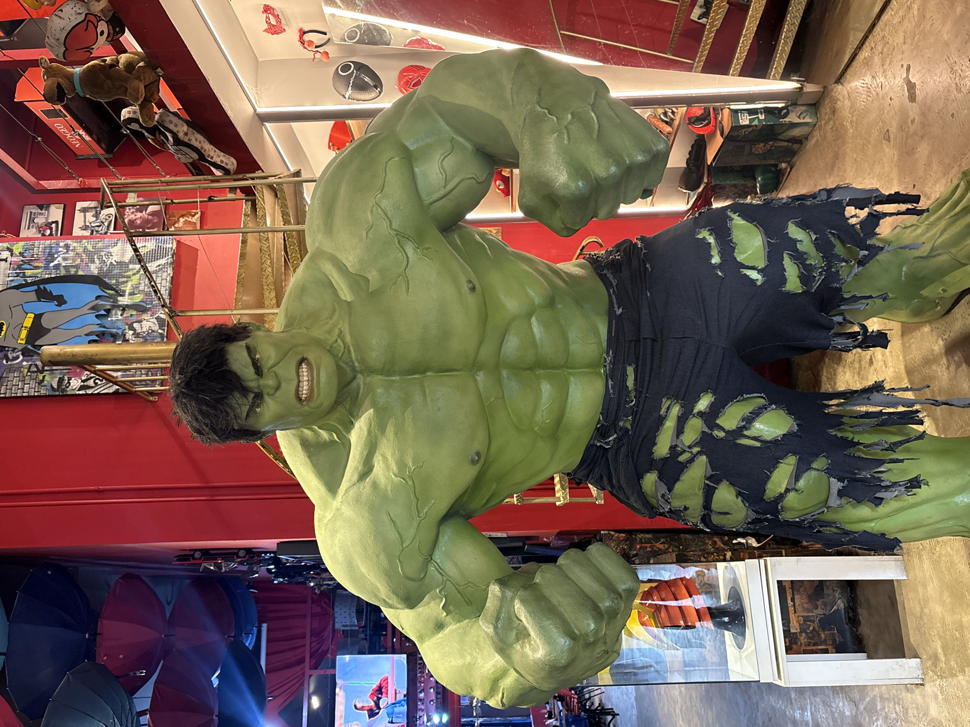 The image displays a life-size statue of the Hulk, a fictional superhero, positioned in the foreground. The statue is predominantly green with visible musculature and is wearing tattered dark pants. Behind the statue, a red wall is visible, adorned with various comic book-style artwork and framed pictures. To the right, a metal spiral staircase ascends, with items like a Scooby-Doo plush toy and other decorative objects placed on its steps and landing. Further to the right, shelves are stocked with various items, including masks and what appear to be novelty items or collectibles. In the background, to the left of the Hulk statue, a display case contains a mannequin wearing an orange and red outfit. The overall environment suggests a retail or entertainment space, possibly a themed store or a pop-culture exhibition. This scene is located at the "Parque da Mônica" in São Paulo, Brazil, a theme park based on the Monica's Gang comic series.