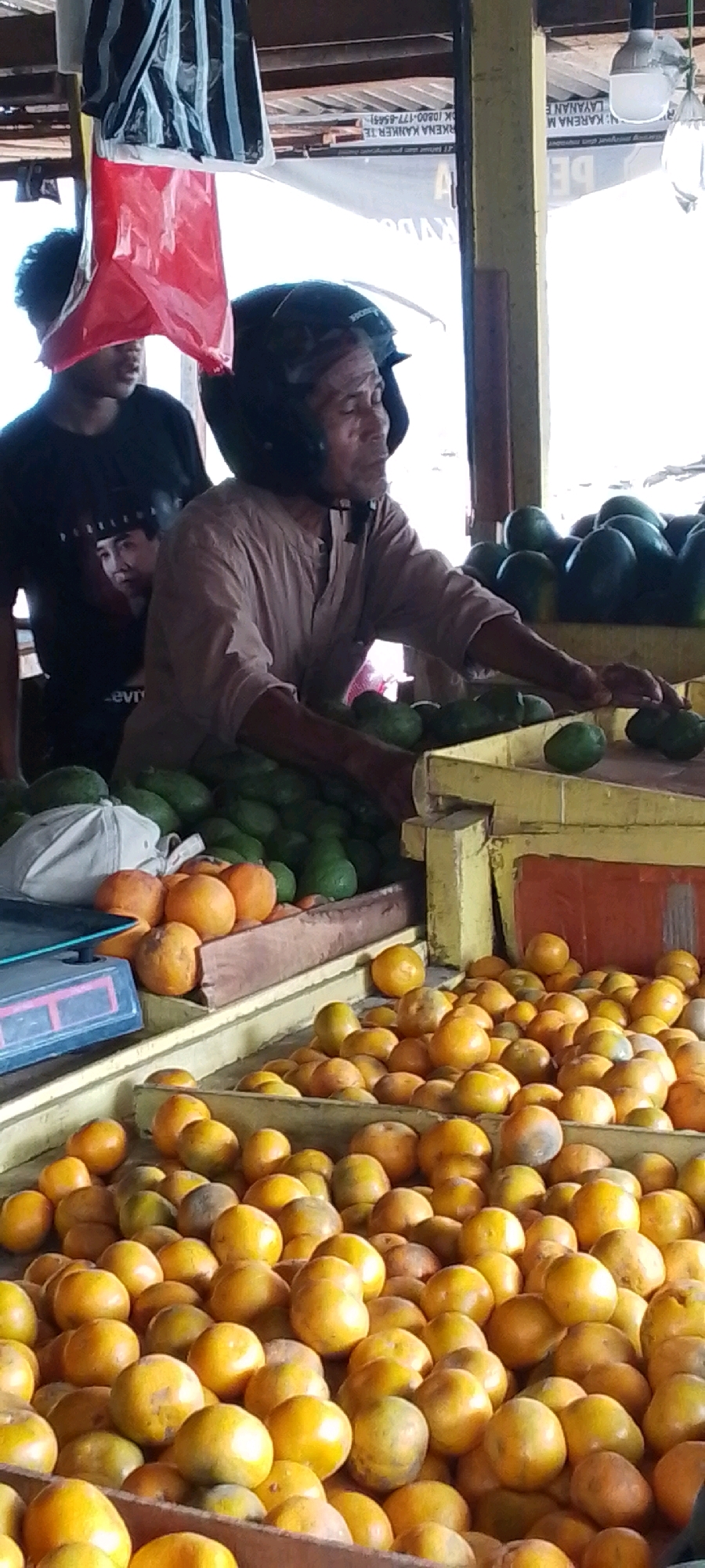 A man wearing a black helmet is standing behind a market stall in Cilaku, Indonesia. He is wearing a light brown, collared shirt and is reaching towards a display of green produce, likely avocados. To his left, another person wearing a black t-shirt with a picture of a man on it is partially visible. In front of the man and the stall, there are large quantities of orange produce, likely oranges or tangerines, piled in wooden crates. Behind the man, a red plastic bag hangs from a support structure. The market appears to be outdoors, with a corrugated metal roof overhead. There is also a scale on the left side of the image.