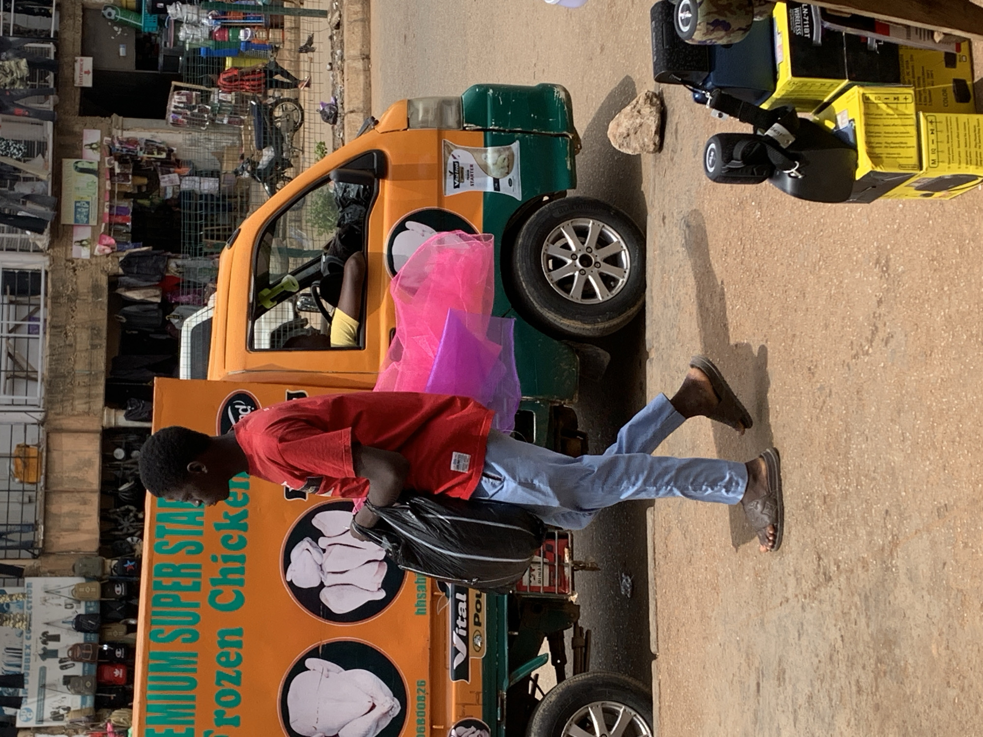 In the city of Bukuru, Nigeria, on a dusty, paved street, a young Black male, appearing to be in his late teens or early twenties, walks from left to right, carrying a large black plastic bag in his right hand and a brightly colored pink garment over his left arm. He is wearing a red t-shirt and light blue trousers, with dark-colored flip-flops on his feet.
Behind him, an orange and green truck is parked. The side of the truck features an advertisement for "PREMIUM SUPER STAR Frozen Chicken" with images of whole chickens. Another advertisement for "Vital Feed" with an image of a chick is visible below the chicken advertisement. The truck's driver's side door is open, and a person wearing a yellow shirt is visible inside the vehicle.
The street is lined with various market stalls and shops. In the background, clothing and accessories are displayed for sale, with racks of garments and hanging bags. A motorcycle is parked to the right of the truck. In the foreground, to the right of the image, several boxes are stacked, topped with black electronic devices, possibly speakers. A large rock sits on the ground near the truck. The overall environment suggests a busy, commercial area.