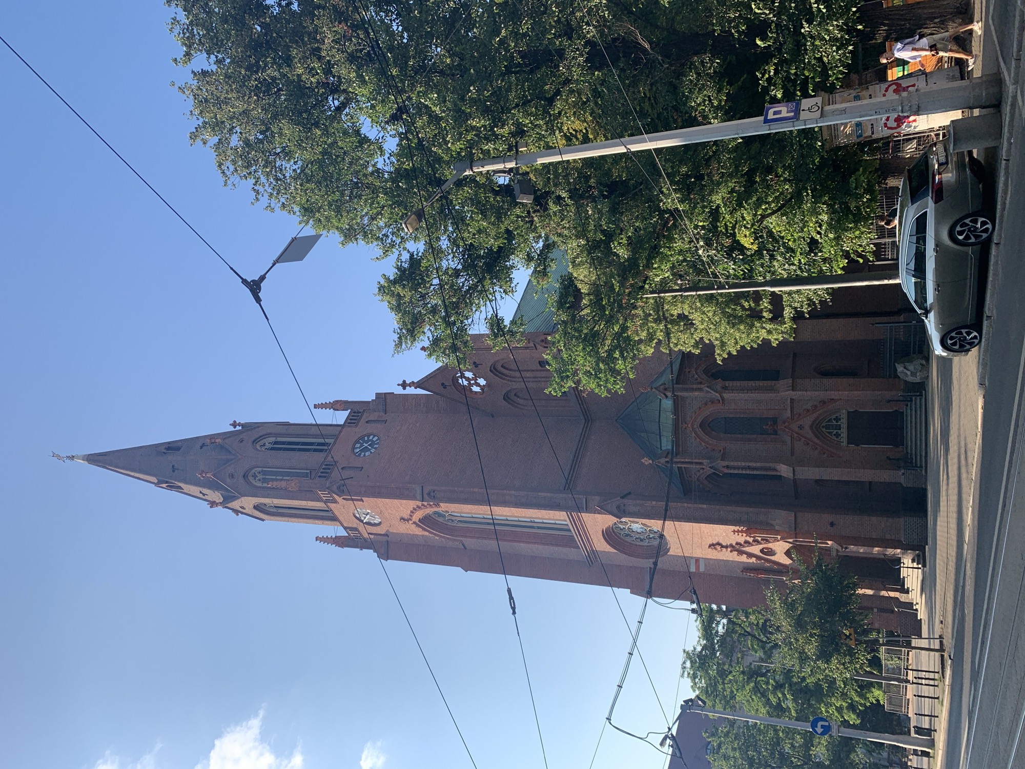 The image shows the Church of the Sacred Heart of Jesus in Poznan, Poland, on a clear day. The red brick Gothic Revival church is tall and imposing, with a prominent spire topped by a cross. A large clock face is visible on the tower. The church is surrounded by trees and there are power lines strung across the sky in the foreground. A modern silver station wagon is parked on the street in front of the church. A few people are visible walking on the sidewalk. The scene appears to be a street view in an urban setting.