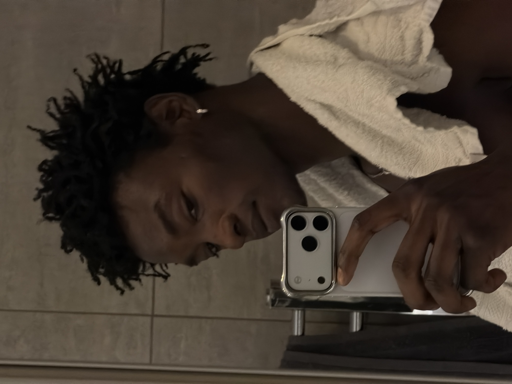 A man with dark, curly hair takes a selfie in what appears to be a bathroom. He is looking at his phone, which is held in his right hand. The phone has a silver case with a triple-lens camera. He is wearing a white towel draped over his shoulders. The background shows tiled walls and a towel rack with dark towels. The lighting is dim. There is no indication of a specific location within the city of London, United Kingdom.