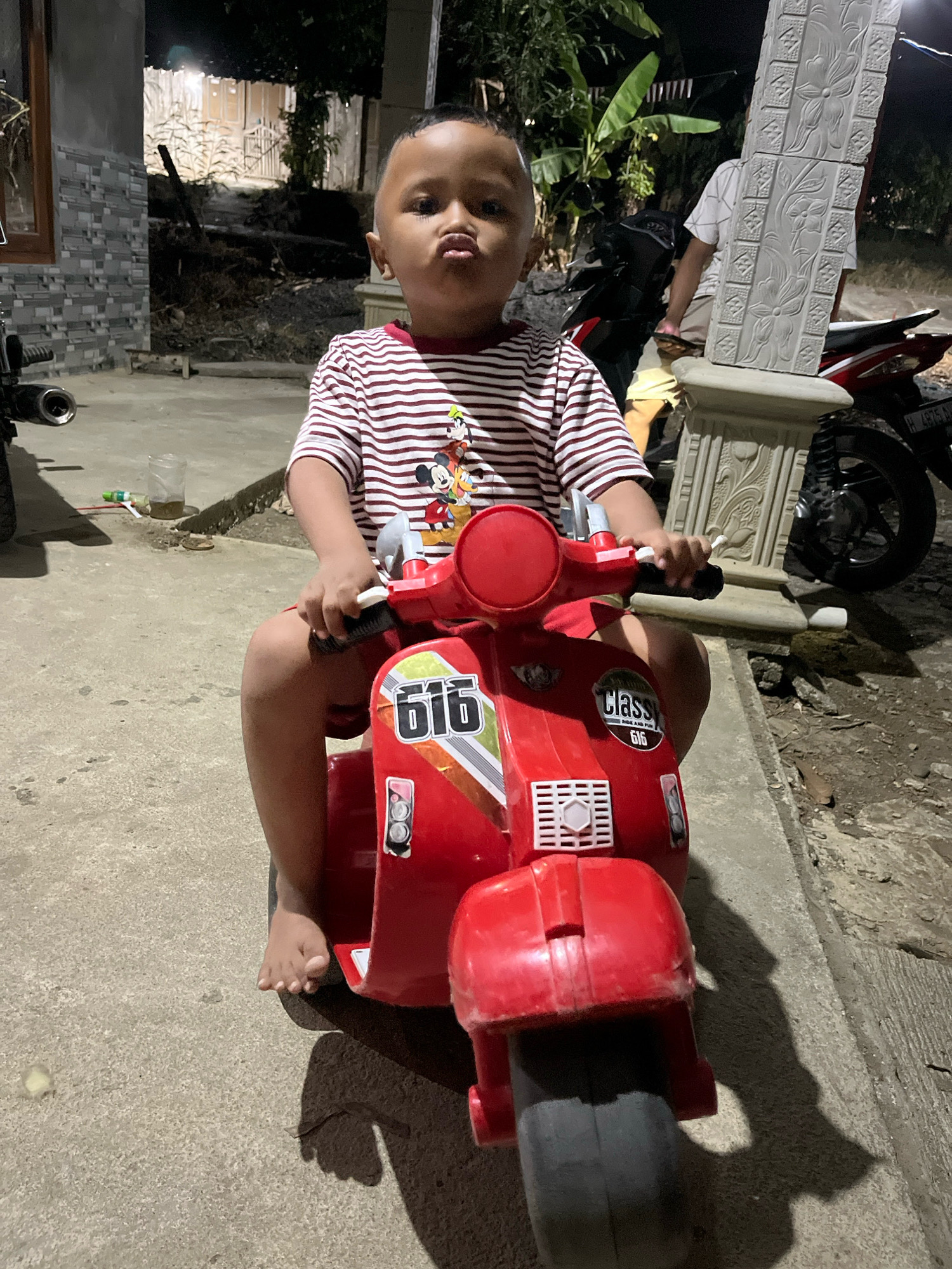 A young child, appearing to be of Southeast Asian descent, sits on a red toy motorcycle. The child is wearing a striped t-shirt with a cartoon character print. Their feet are bare and touch the ground. The motorcycle is parked on a paved surface, possibly a driveway or sidewalk. In the background, there are structures that suggest a residential area, with some vegetation visible. A portion of a decorative pillar with floral carvings stands to the right of the child. A second motorcycle is partially visible behind the pillar. The lighting suggests it is evening or nighttime. The scene appears to be in Wonokerto, Indonesia, as indicated by the context.