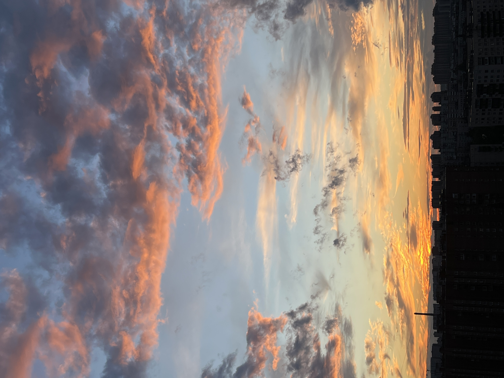 The image depicts a panoramic view of a cityscape at sunset, likely Sinyavino, Russia. Silhouetted apartment buildings form a dense urban horizon against a dramatic sky. The clouds are illuminated with vibrant hues of orange, pink, and yellow from the setting sun, contrasting with patches of dark grey and pale blue in the sky. A tall, slender industrial chimney is visible amongst the buildings. The overall scene is one of urban development under a colorful and dynamic sky.