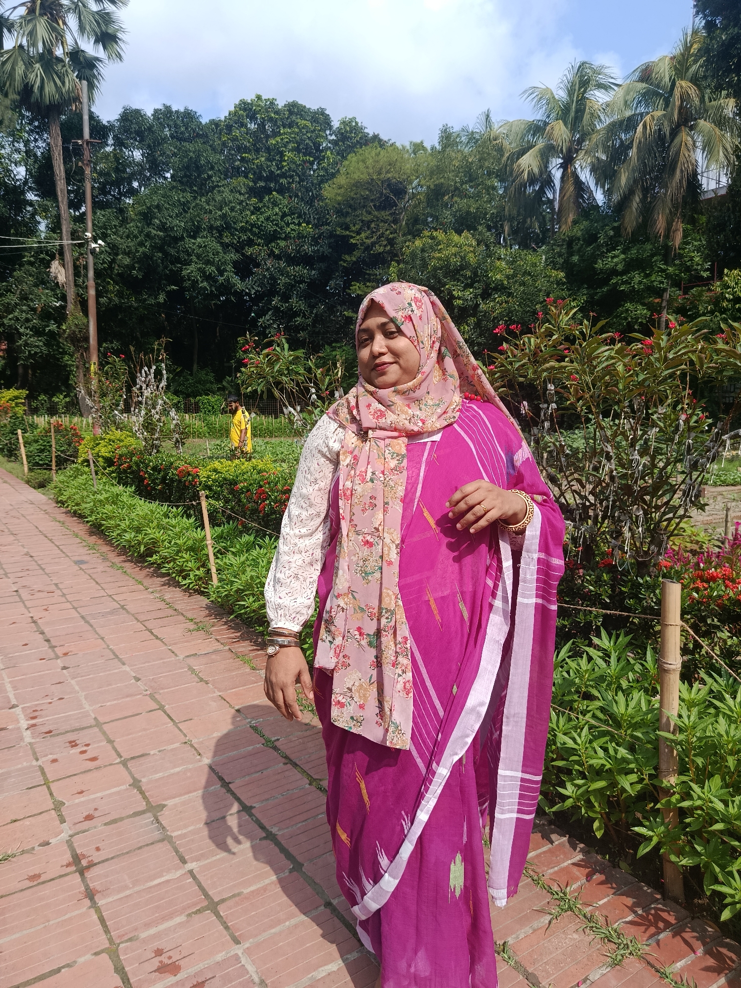 A woman stands on a brick path in a garden, likely in Dhaka, Bangladesh. She wears a magenta sari with white stripes, a long-sleeved white blouse with lace detailing, and a floral-patterned headscarf. The path is lined with plants and flowers, with taller trees and shrubs in the background. A figure in a yellow shirt is visible in the distance. Palm trees and utility poles are also present in the background, further suggesting a park setting.
