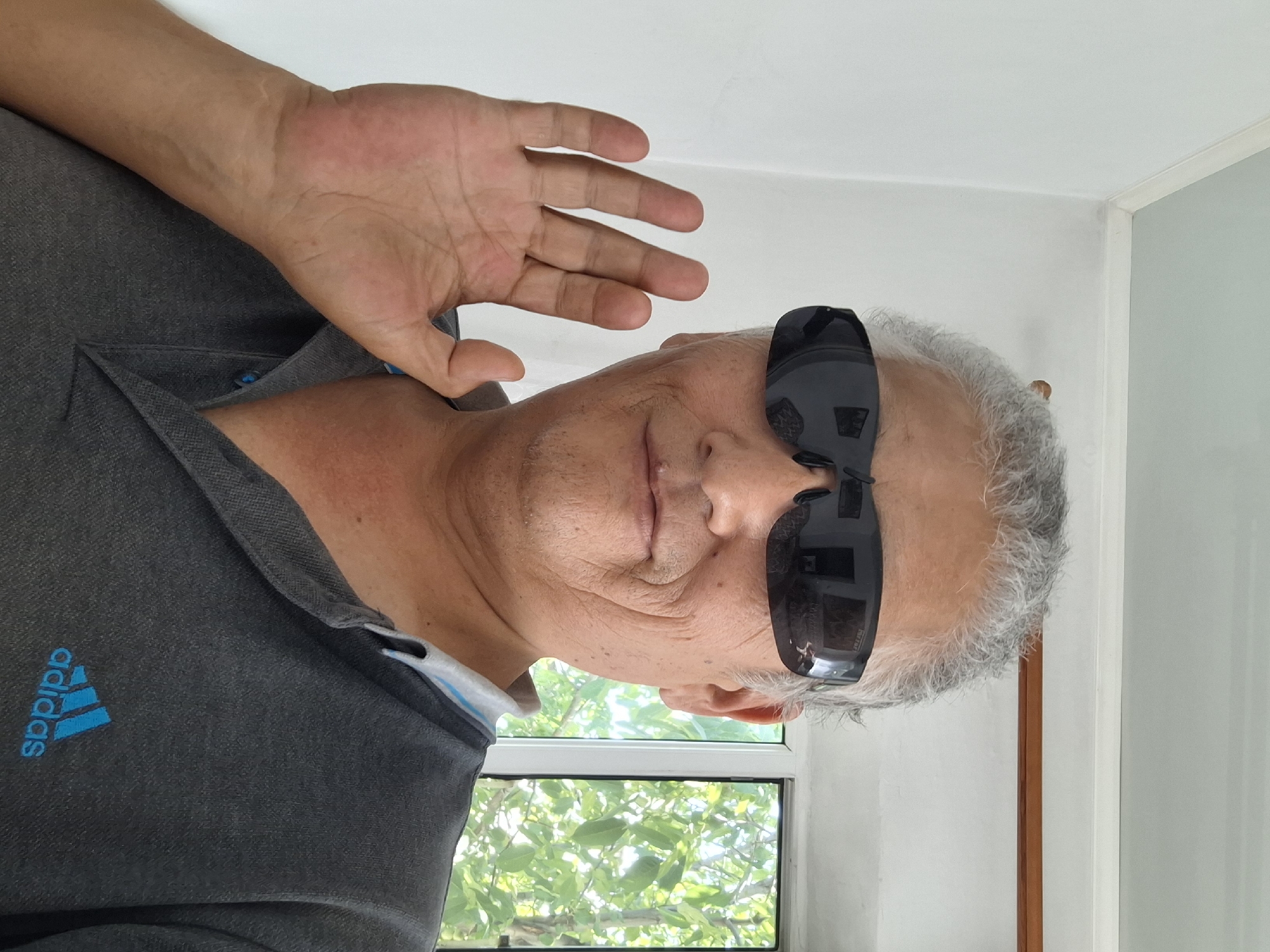 A medium shot shows a man with graying hair and sunglasses, wearing a dark gray Adidas polo shirt. He is holding his left hand up, palm facing the camera, as if waving. The background consists of a white wall with a blue ceiling and a window revealing green trees outside. The image appears to be a selfie, taken indoors. The specific location in Almaty, Kazakhstan, is not discernible from the image.