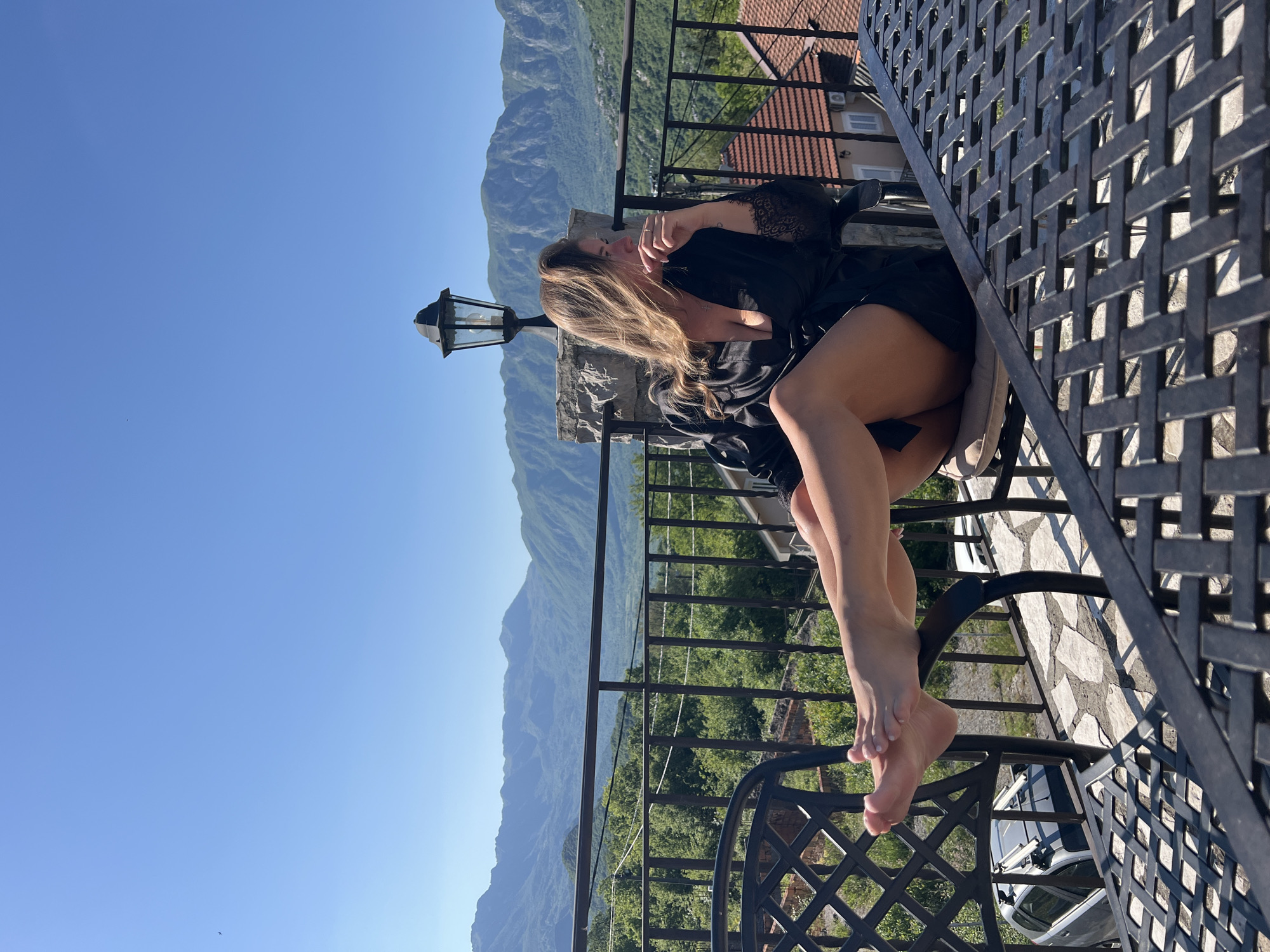 A woman in a black robe sits on a black wrought iron chair on a balcony overlooking a mountainous landscape under a clear blue sky. A black wrought iron table sits beside her. A street lamp stands next to the balcony. The background includes buildings with red tile roofs, trees, and mountains. The location is likely Sotonići, Montenegro.