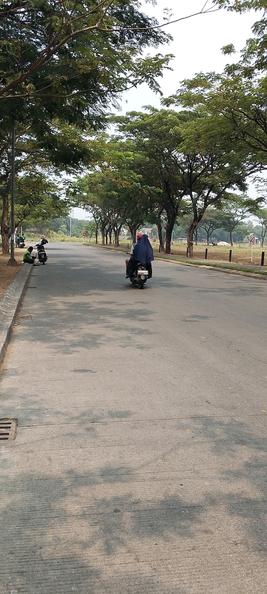 The image depicts a road in Bekasi, Indonesia, lined with trees casting shadows on the asphalt. In the foreground on the left, two individuals are seated on the curb next to a parked motorcycle. Further down the road, a motorcycle carrying two passengers, one wearing a blue hijab, is moving away from the camera. The background shows a park-like area with more trees and clear sky.