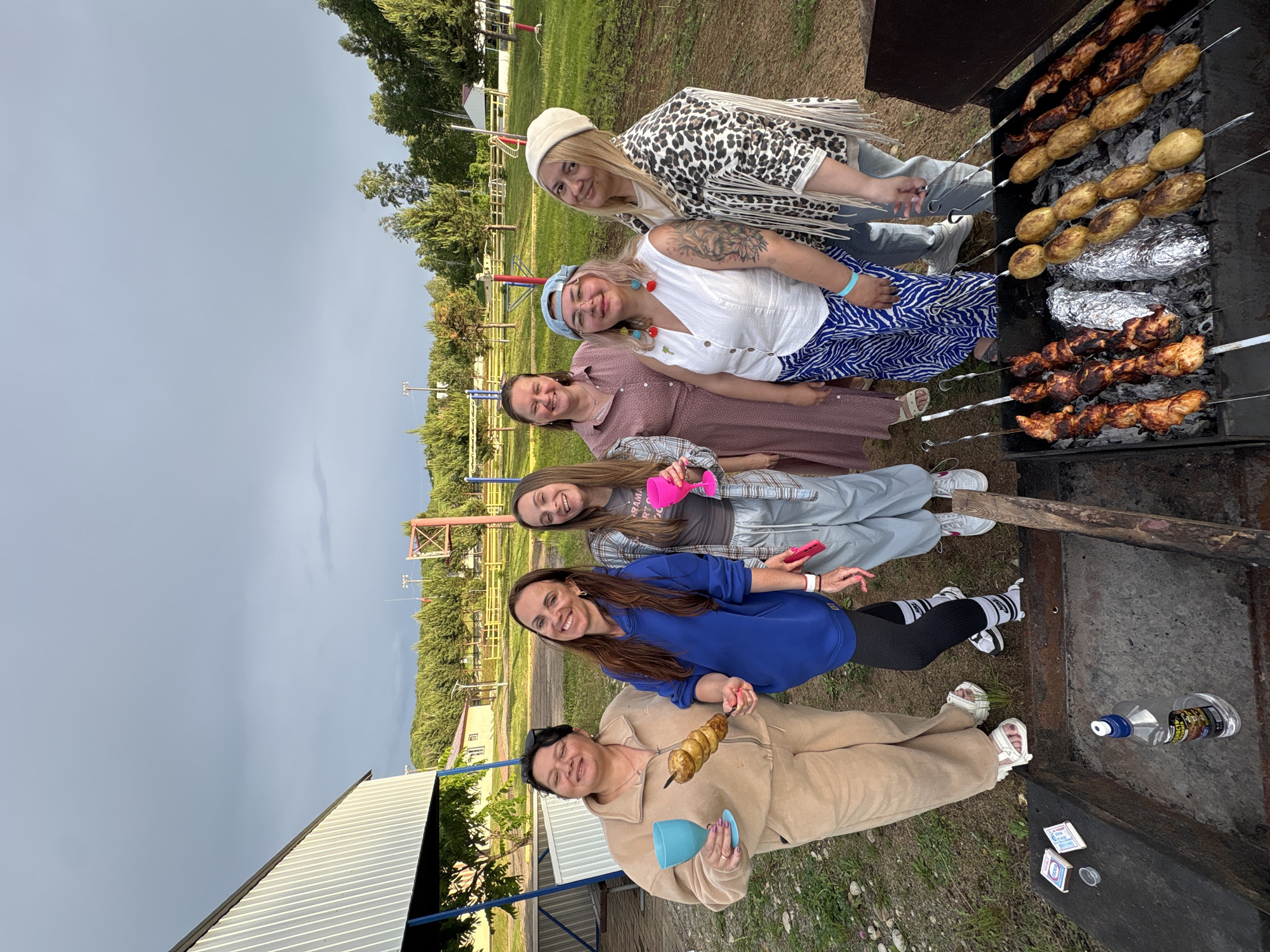 The image depicts a group of six women outdoors, seemingly at a picnic or barbecue. They are standing around a barbecue grill laden with skewers of meat and potatoes wrapped in foil. In the foreground, on the edge of the grill, are matches and a bottle of alcohol. The women are smiling and appear to be enjoying themselves.
The background reveals a park-like setting with trees, a playground structure, and a building that resembles a small pavilion or shed. The sky is overcast with dark clouds, suggesting an impending storm.
The attire of the women varies, ranging from casual tracksuits and sweatpants to dresses and patterned jackets. Some are holding drinks. The overall atmosphere is relaxed and social.
Based on the presence of the specific brand of alcohol and the type of grilled food, along with the general ambiance, this scene is consistent with a gathering in a recreational area within or on the outskirts of Birobidzhan, Russia. The setting appears to be a public park or a private recreational plot, common in such locations for outdoor activities.