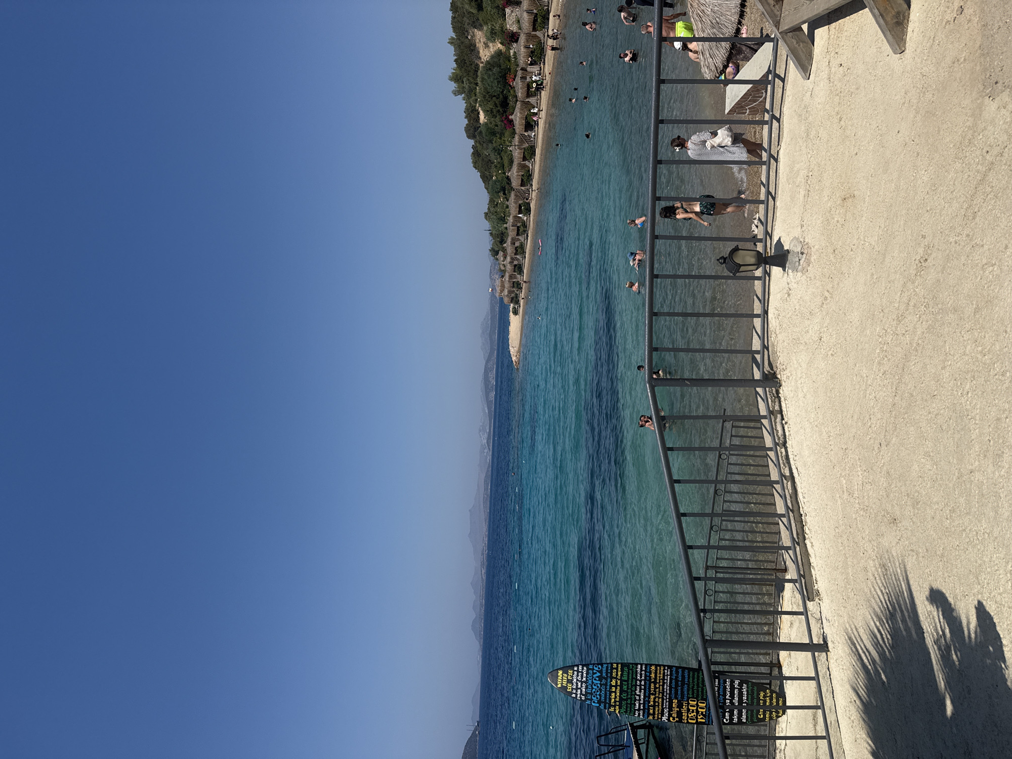 The image depicts a picturesque beach scene in Bodrum, Turkey, on a clear, sunny day. The foreground features a sandy area with a grey metal railing. Beyond the railing, a body of crystal-clear turquoise water beckons, with numerous people swimming and enjoying the sea. In the middle ground, a sandy shoreline is dotted with numerous thatched-roof cabanas, suggesting a resort or beach club setting. Lush green trees and vegetation line the hills in the background, with faint outlines of mountains visible on the horizon. A large, dark sign with white text is partially submerged in the water near the left edge of the frame, providing information or rules for the beachgoers. The overall atmosphere is one of leisure and relaxation, characteristic of a popular coastal destination.