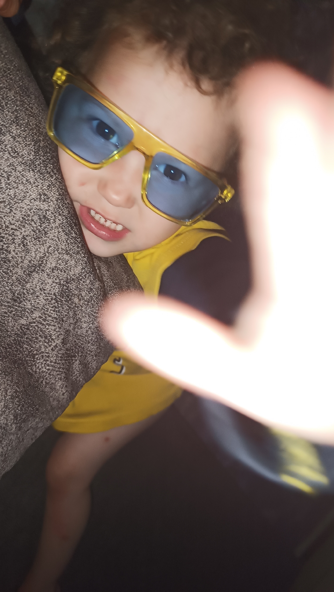 A close-up, vertical, low-angle shot captures a young child wearing oversized yellow-framed sunglasses with blue lenses. The child's face is partially obscured by a hand, and their curly brown hair is visible at the top of the frame. The child appears to be looking at the camera, with their mouth open slightly, revealing teeth and pink lips. They are wearing a yellow shirt with black accents. The child is leaning against a textured, brown surface, possibly a couch or a wall. The background is dark and out of focus, suggesting an indoor setting. There is no discernible information about the location in Karsha, Russia.
