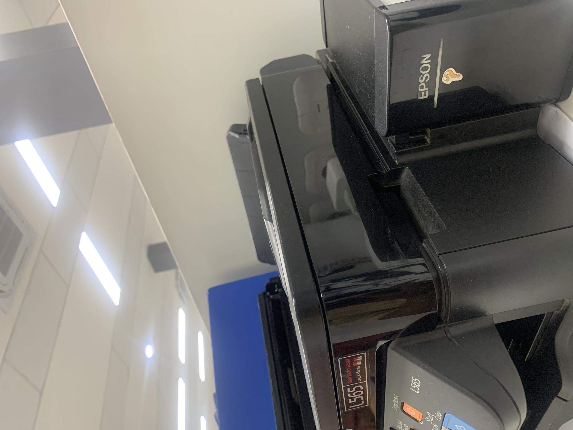 The image shows the top portion of a black Epson printer, model L565, with a blue partition visible in the background. The printer is situated in what appears to be an office environment, with overhead fluorescent lighting and a tiled ceiling. A sticker of a cartoon question mark is affixed to the side of the printer. The scene is clean and well-lit, suggesting a typical office setting in Palembang, Indonesia.