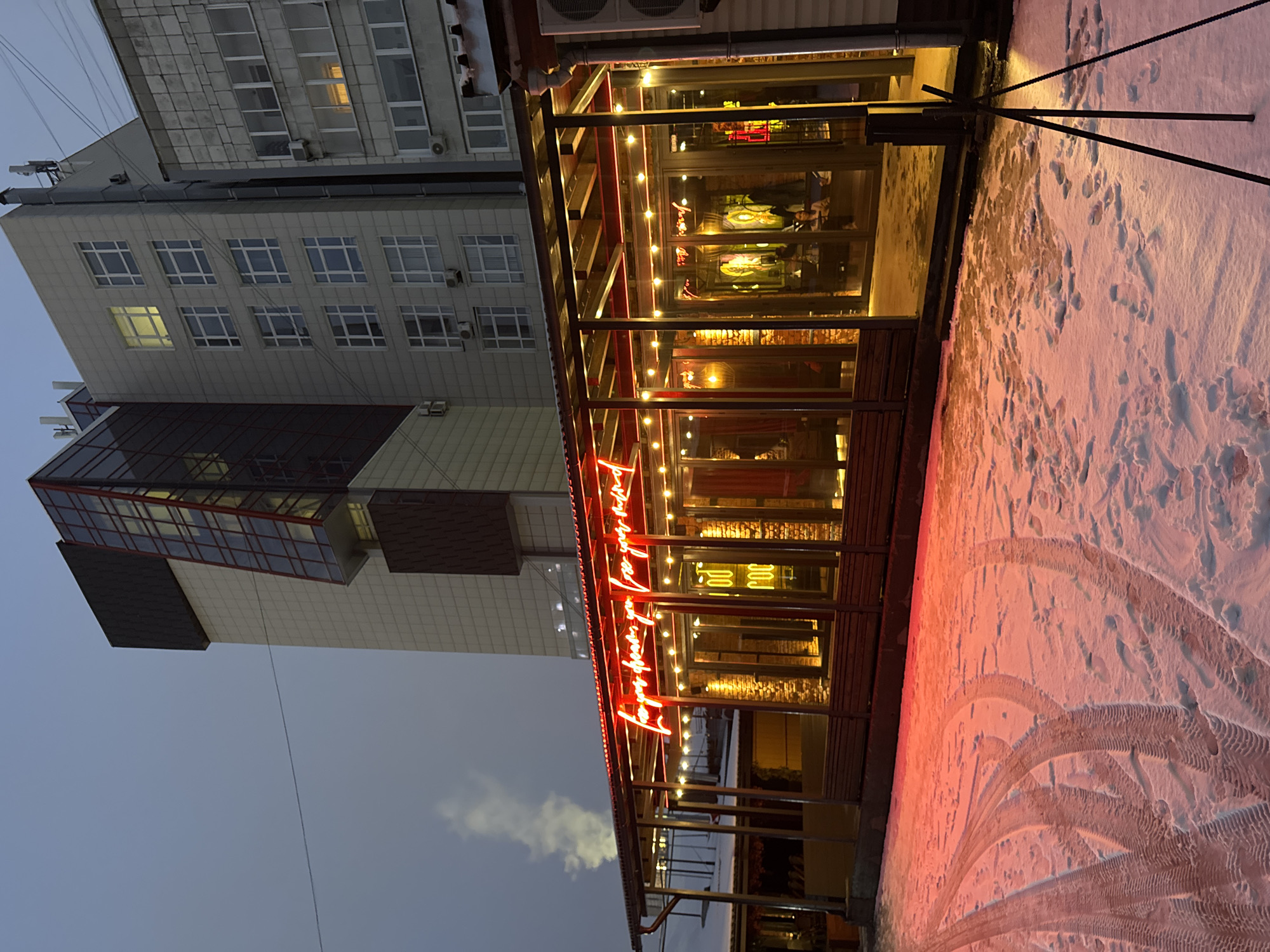 The image captures an evening or late dusk scene in Perm, Russia, showcasing a modern urban environment during winter. On the left, a multi-story building with a light-colored facade features numerous square windows, some of which are illuminated, casting a warm yellow glow. The building exhibits a contemporary design with a prominent dark-paneled, possibly glass, section protruding at a lower level. Air conditioning units are visible on its exterior.
To the right, a glass-enclosed outdoor patio or restaurant extends from a building, serving as a focal point. This structure is brightly lit with numerous warm yellow string lights and a prominent red neon sign, which displays cursive English text that is partially discernible, possibly reading "Love you for all...". Inside the enclosed space, faint figures of people can be seen, suggesting they are dining or socializing. The interior also shows tables, chairs, and brickwork.
The ground in front of the patio is covered in a layer of snow, tinted reddish by the glow of the neon sign. Clear tire tracks and various footprints are imprinted on the snow, indicating recent activity. A dark, multi-legged support structure is visible on the snow-covered ground near the patio. The sky above is a dark, overcast blue-grey. The warm, inviting light of the patio creates a stark and cozy contrast against the cold, snowy exterior and the muted tones of the sky and background building, suggesting a vibrant spot in a chilly environment.