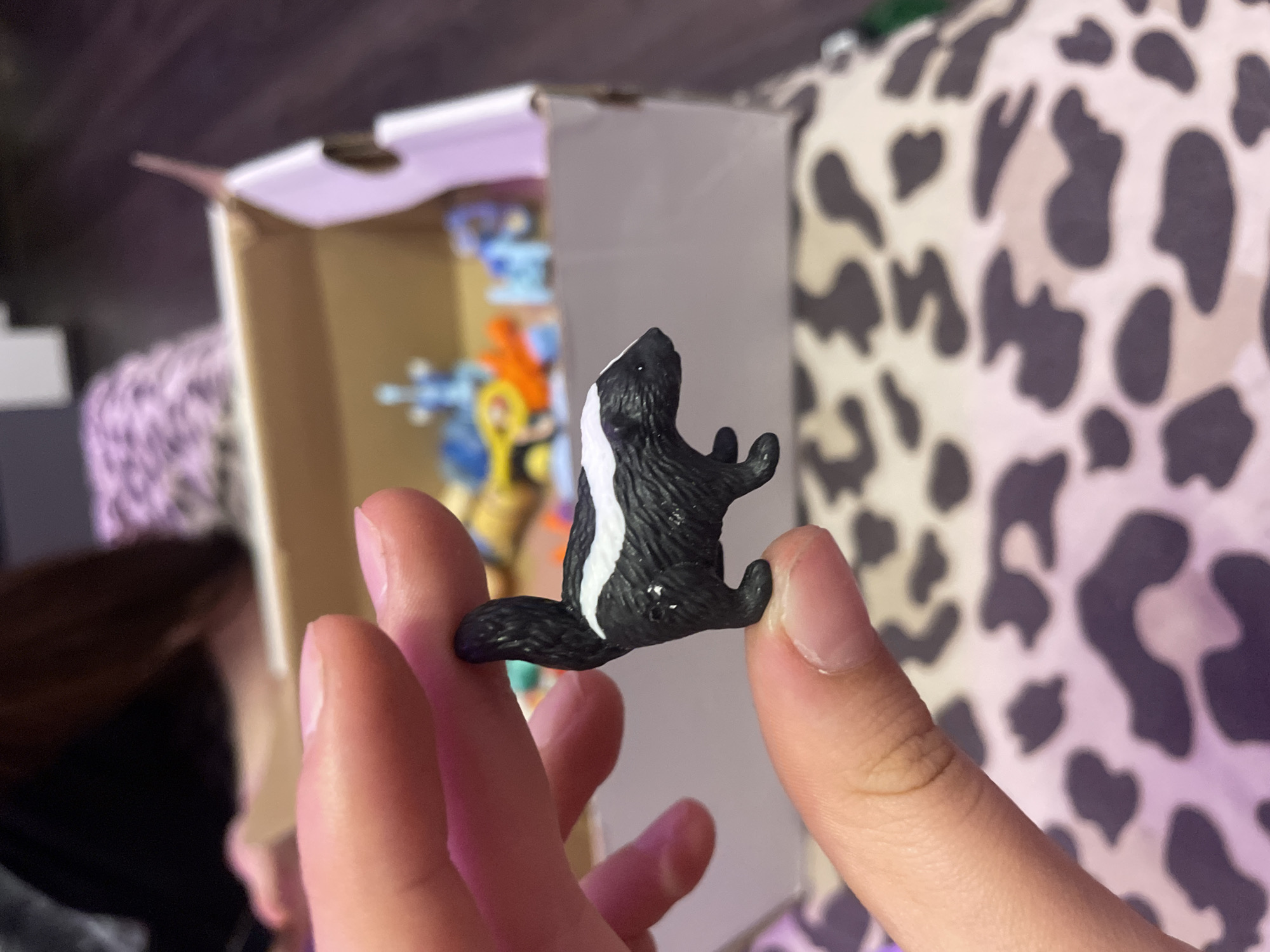 A person's fingers hold a small, black and white plastic toy skunk. The skunk is detailed with a white stripe running down its back and bushy tail. In the background, a cardboard box contains other blurred plastic toys. The scene appears to be indoors, with a person's dark hair and a purple and white patterned surface visible. The location is Ladozhskii Trudposelok, Russia.