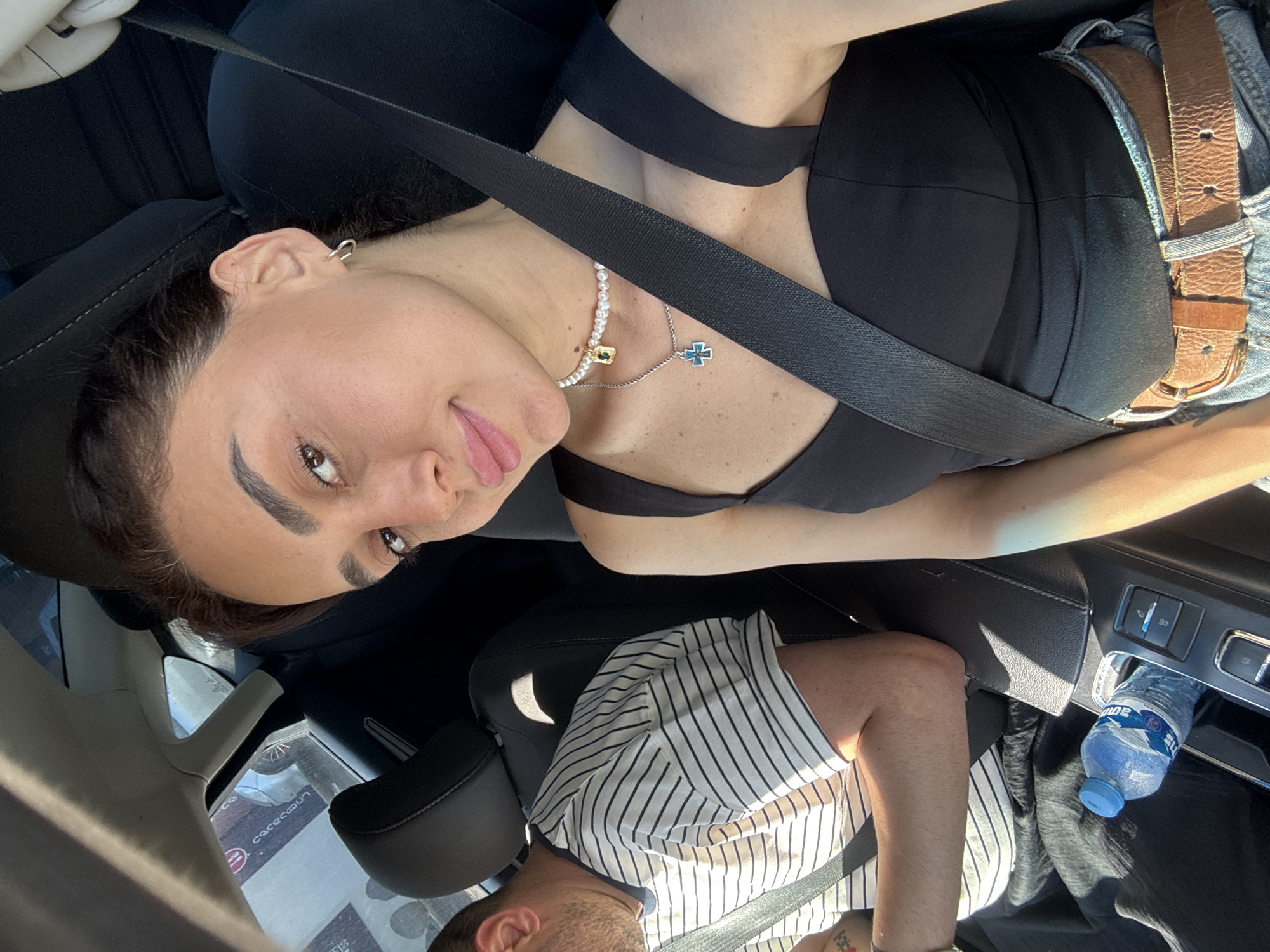 This is a close-up, selfie-style photograph taken inside a car, likely in Tbilisi, Georgia. The main subject is a woman in the passenger seat, looking at the camera with a slight smile. She is wearing a black top, blue jeans, and two necklaces, one with pearls and a pendant, and another with a cross pendant. She has a seatbelt fastened across her body. In the background, a man is visible in the driver's seat, wearing a striped t-shirt and also wearing a seatbelt. A bottle of water is placed in the center console of the car. The interior of the car appears to be modern, with a black leather trim and dashboard. Sunlight is streaming into the car, creating shadows and highlights on the subjects and interior. The overall impression is of a casual moment captured during a car journey, with hints of the urban environment visible through the car windows.