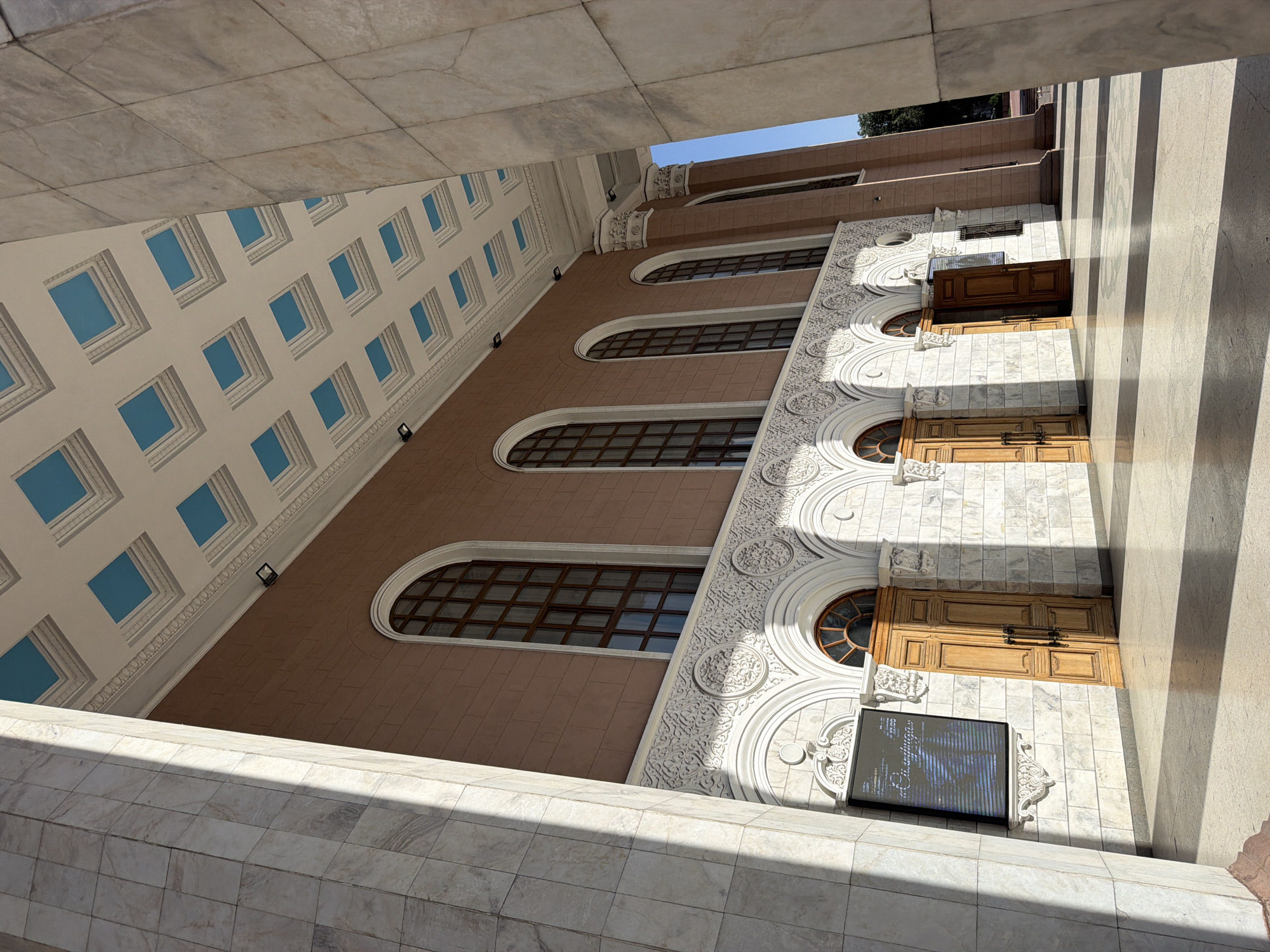 A low-angle shot captures the exterior of a building in Bishkek, Kyrgyzstan. The building features a symmetrical design with a section of arched windows and an adjacent section with a grid of square, recessed window openings. The building materials include marble or a similar stone, which forms the visible framing structures. The architecture is intricate, with detailed ornamentation around the arches and windows.
Three wooden doors are visible, each framed by decorative stone elements. A sign with lettering is located near one of the doors. The lighting is bright and casts shadows, emphasizing the texture and depth of the building's facade. The perspective highlights the height of the building and the surrounding architectural elements.