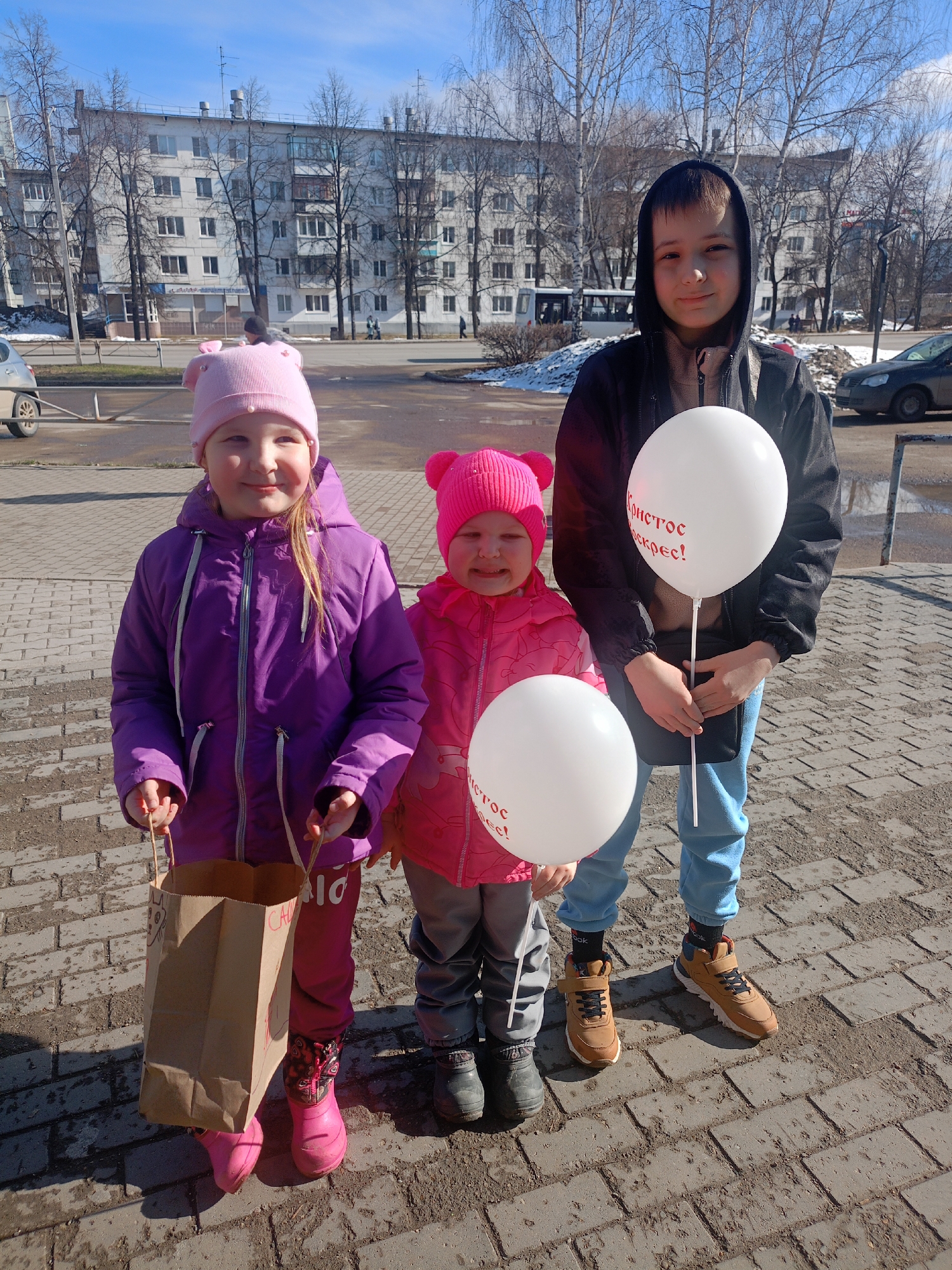 Three children stand on a paved area with a large building in the background. The girl on the left wears a purple jacket, pink pants, and a pink hat with decorative ears, carrying a brown paper bag. The girl in the middle is dressed in a pink jacket, gray pants, and a pink hat, and she holds a white balloon. The boy on the right wears a dark jacket, light blue pants, and holds another white balloon. Text in red is visible on the balloons. A street with cars and leafless trees completes the scene. The location is identified as a city in Unknown, Unknown.
