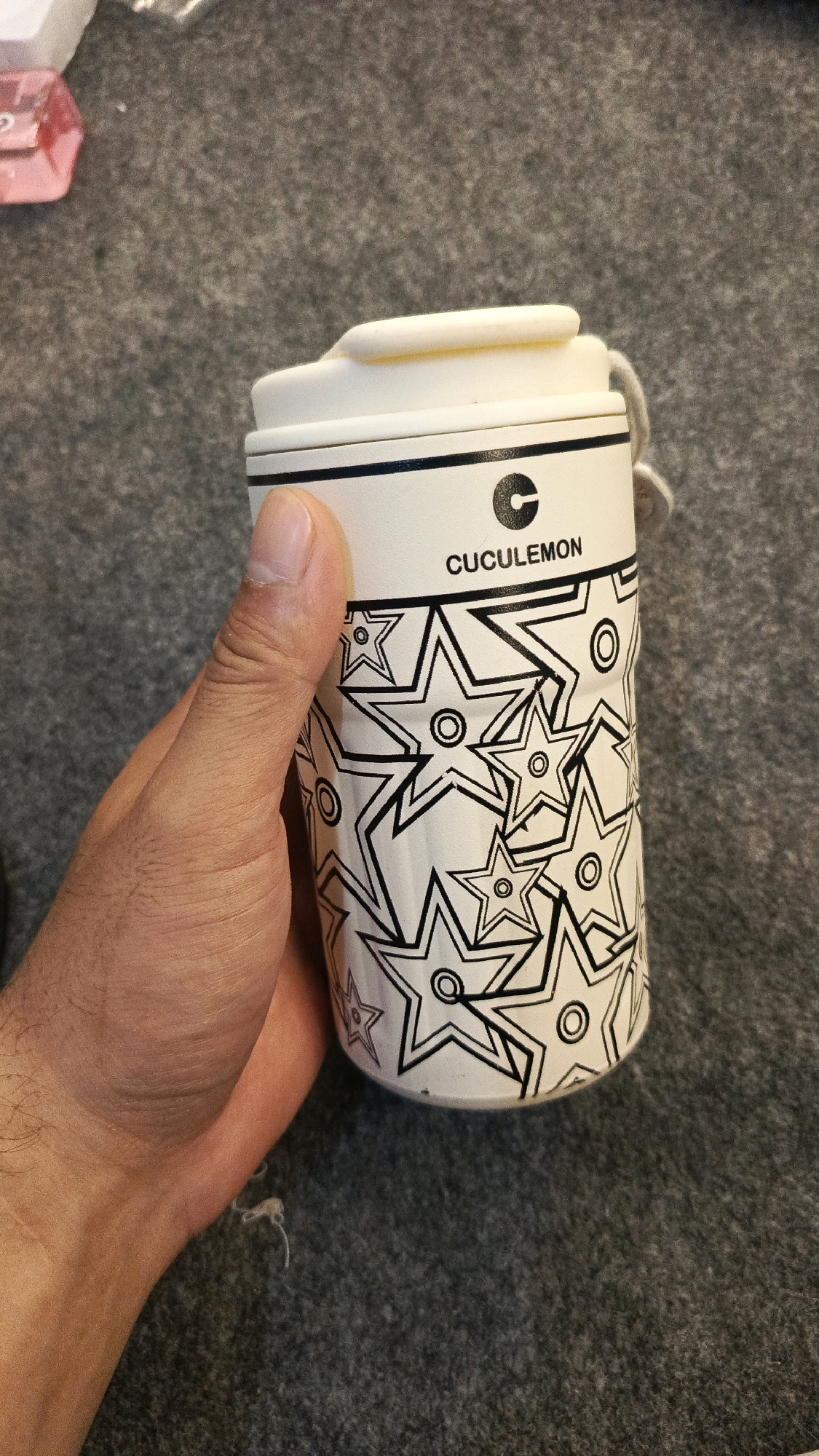 A hand holds a white cylindrical cup with a black star pattern and the word "CUCULEMON" printed on it. The cup has a white lid with a loop. The background is a textured gray surface, possibly a carpet or fabric. There are no visible elements that would indicate a specific location in Babadan, Indonesia.
