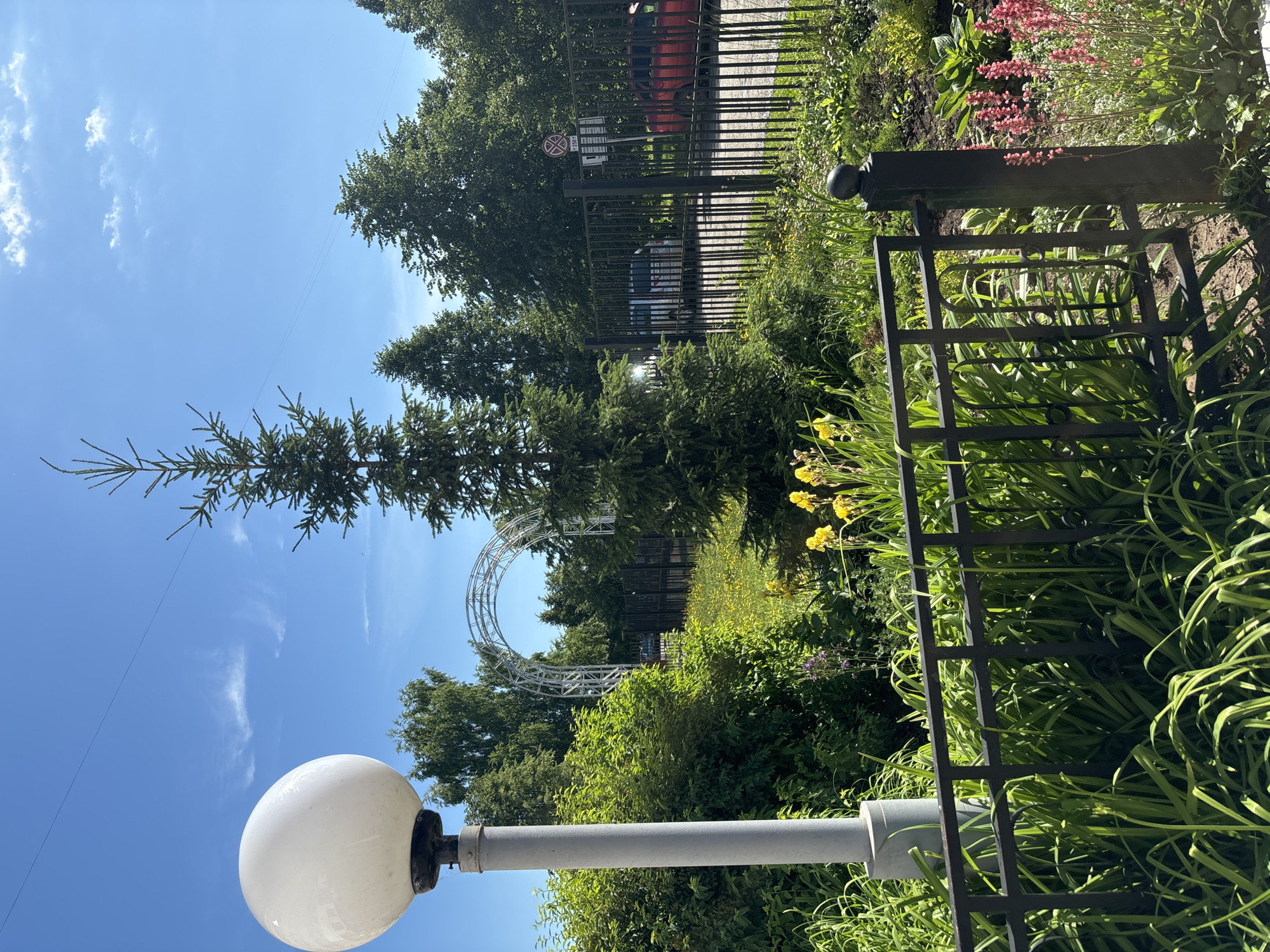 The image shows a street view in Chaikovsky, Russia. In the foreground, a black metal fence encloses a garden with lush green plants and some yellow flowers. To the left of the fence, a tall, grey lamppost with a white spherical light fixture stands prominently. Behind the fence, a black metal gate separates the garden from a street where two cars are visible, one red and one silver. A traffic sign is mounted on a pole near the gate. In the middle ground, a large evergreen tree is situated, and beyond it, a white arched structure can be seen. The background consists of dense green trees under a clear blue sky with a few wispy clouds. The scene appears to be set during the daytime with bright sunlight.