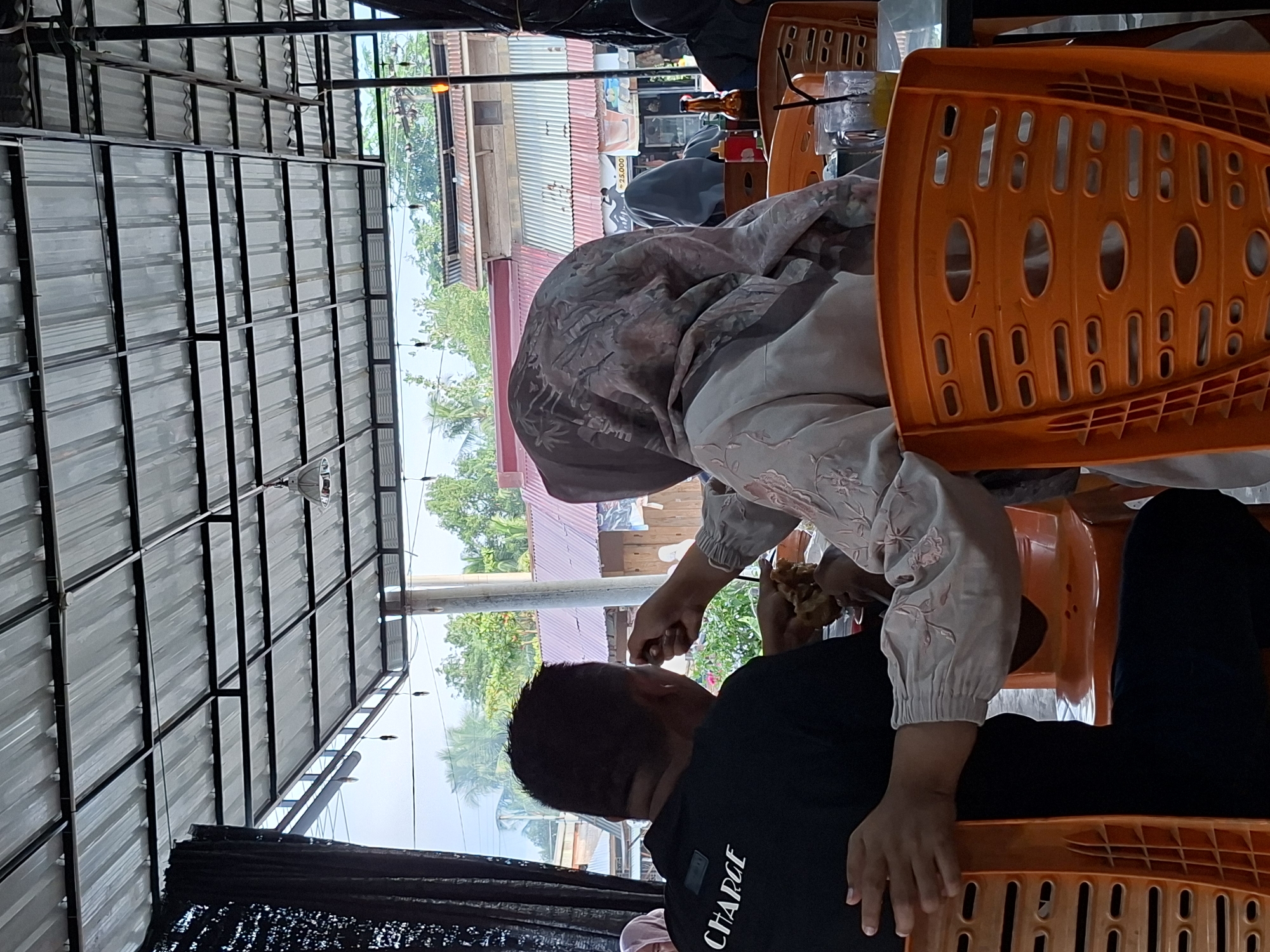 A person wearing a headscarf and a light-colored patterned blouse is feeding another person sitting across from them. The person being fed is wearing a black shirt with the word "CHARGE" printed on it. They are seated on orange plastic chairs under a metal awning. In the background, buildings and trees are visible. The scene likely takes place in a restaurant or cafe. The location may be Lhokseumawe, Indonesia.