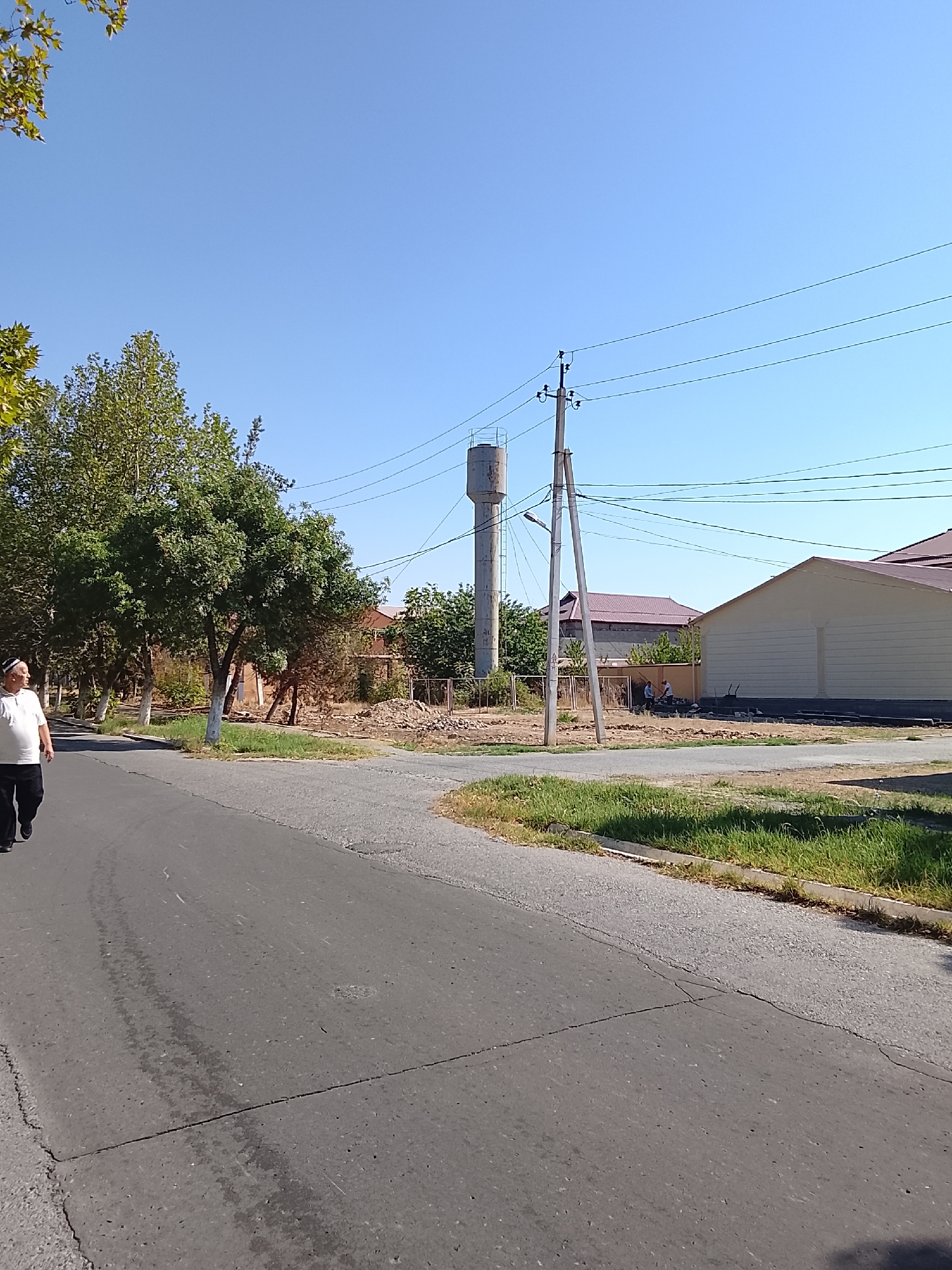 In Pakana, Uzbekistan, a man wearing a white polo shirt and dark pants walks along a cracked asphalt road. To his left, trees line the roadside, their leaves green under a clear blue sky. In the background, a concrete water tower stands tall, accompanied by a utility pole supporting electrical wires. Several buildings with reddish-brown roofs are visible, and a new, lighter-colored building with a textured facade is under construction. Construction workers can be seen in the distance near the construction site. The overall environment appears to be a semi-urban setting with ongoing development.