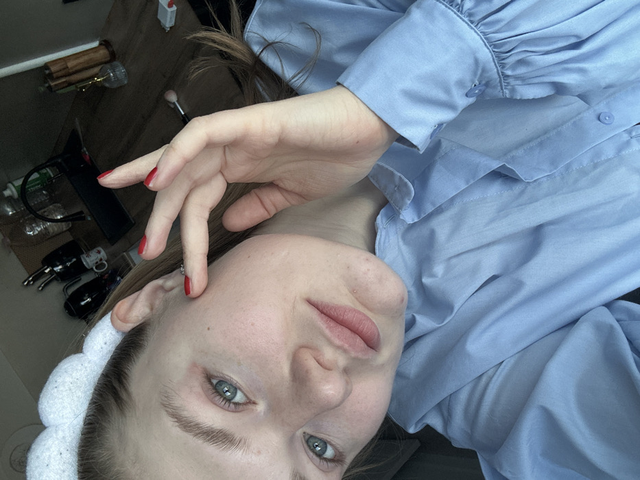 A fair-skinned person with blue eyes and light brown hair wearing a white headband and a light blue button-up shirt is seen in a close-up, slightly tilted view. Red nail polish adorns the fingers of their hand which rests near their face. In the background, a blurred surface holds various objects, including makeup brushes, bottles, and small black devices.