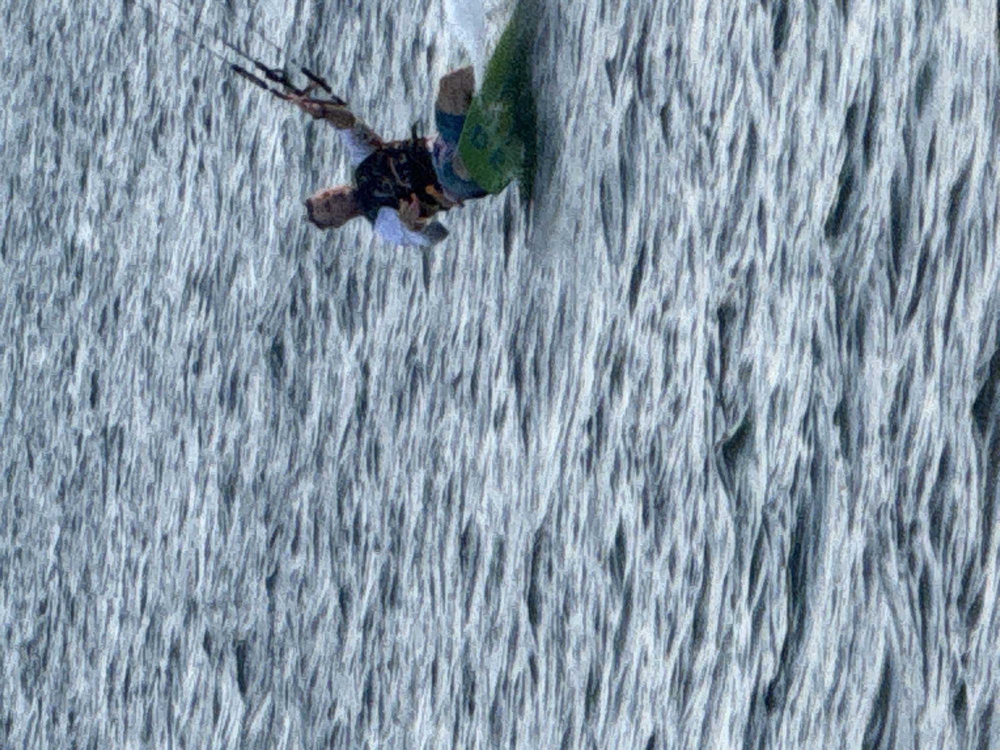 A single person is kite surfing on a body of water. The person is wearing a white shirt, a black life vest, and patterned shorts. They are holding onto a kite bar, which is attached to a kite (not fully visible). The person is positioned on a green surfboard, with white water splashing around them. The background consists of choppy, grey-blue water. The scene appears to be outdoors, on a body of water, possibly the ocean or a large lake, given the waves and open expanse. There is no indication of the city of Denpasar, Indonesia within the image.