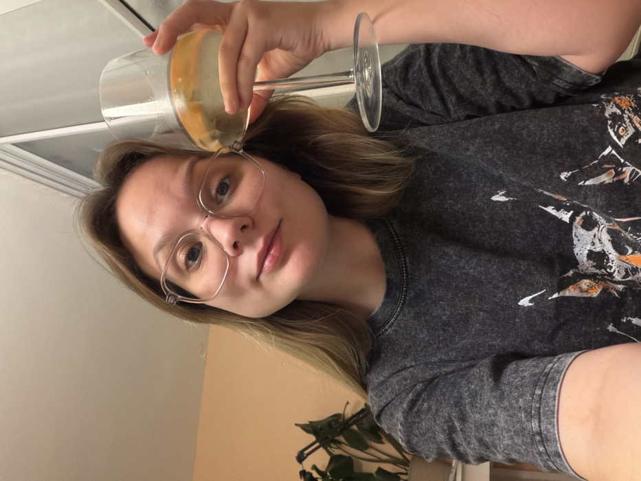 A single female subject is depicted in a selfie-style photograph, taken indoors within a residence located in Tel-Aviv, Israel. The subject, a light-skinned young adult with light brown hair, wears clear-framed eyeglasses. Her attire includes a dark grey, acid-washed t-shirt with a graphic of a stylized animal head, possibly a dog, featuring white and orange accents on the front. She holds a clear stemmed wine glass in her right hand, containing a pale yellow liquid, ice, and several slices of citrus fruit. Her gaze is directed towards the camera, and her lips display a subtle, closed-mouth smile. The background consists of a light peach-colored wall, with a dark green potted plant visible on the left side, and a section of a white window or door frame on the right.