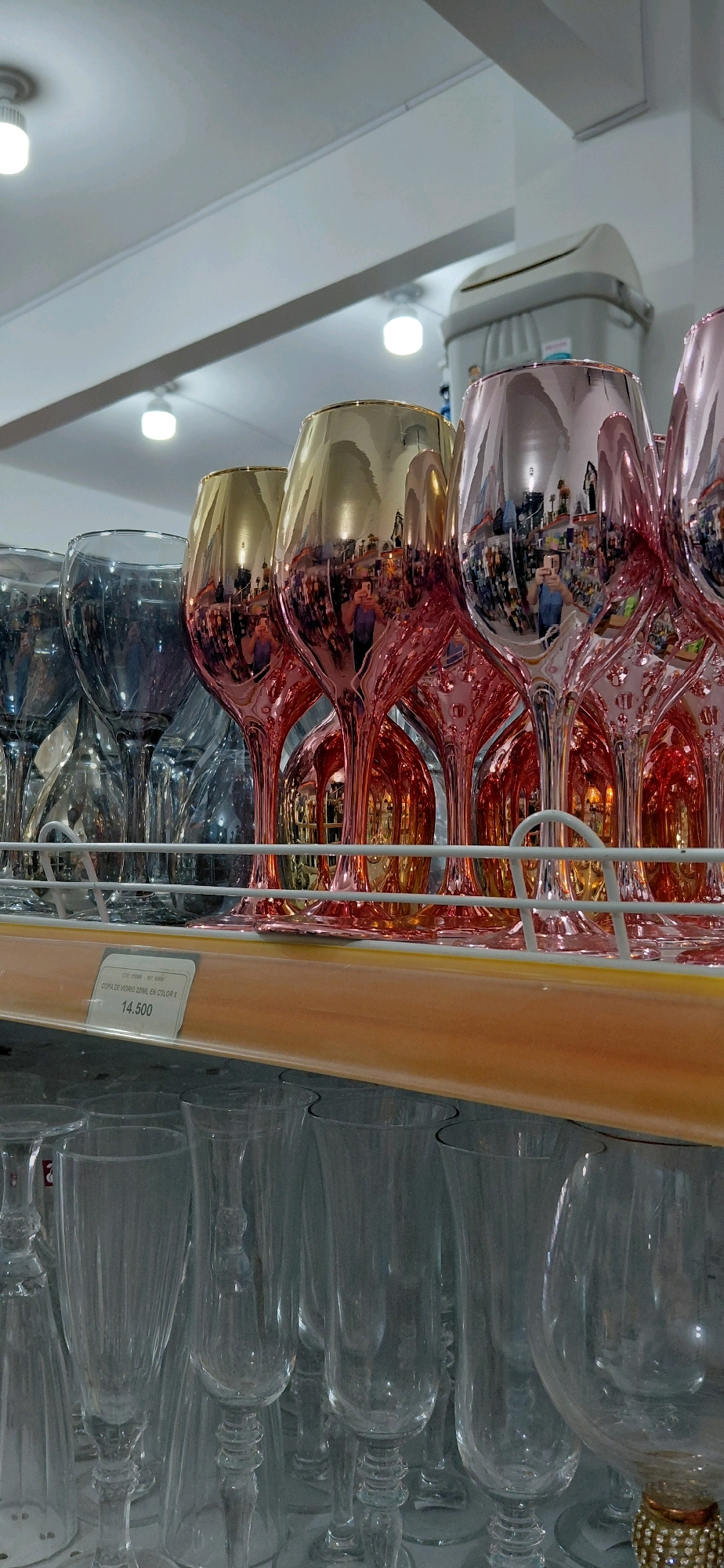 A close-up, eye-level shot captures a retail display of glassware, likely in a store in Encarnacion, Paraguay. In the foreground, a shelf is laden with various wine and champagne glasses. The upper portion of the shelf features a row of iridescent wine glasses in shades of gold and pink, reflecting a brightly lit store interior and a person holding a smartphone, presumably taking a photograph. Behind these, a selection of smokey grey glasses is visible. Below this shelf, another tier showcases a collection of clear, stemmed glasses, some with ornate bases. A price tag, indicating a price of "14.500" and identifying the item as "COPA DE VIDRIO 22ML EN COLOR X", is affixed to the edge of the upper shelf. The background is blurred, showing ceiling lights and shelving units stocked with more merchandise. The overall impression is one of organized retail presentation.