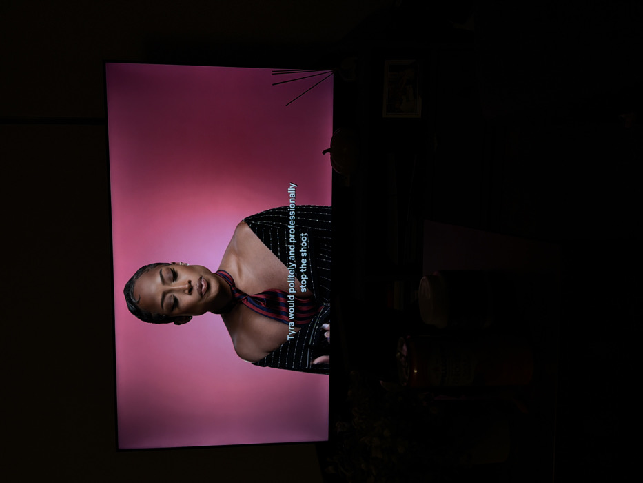 The image displays a dimly lit interior scene in New York, United States of America, focusing on a television screen. The television is centrally positioned, exhibiting a medium shot of a Black female individual against a solid magenta-pink background. She presents with dark, wavy hair styled away from her face, an off-the-shoulder dark striped garment, and a red and navy striped necktie worn loosely around her neck. Her expression is calm, with eyes slightly downcast. White closed captions are visible at the bottom of the screen, stating: "Tyra would politely and professionally stop the shoot". Below the television, on a dark surface, are several objects: a dark leafy arrangement on the left, followed by a beverage can with a visible red label, a light-colored disposable cup with a lid, a small orange decorative pumpkin, a reed diffuser with dark reeds, and a small framed picture on the right. The overall environment of the room is dark, suggesting low ambient lighting, with the television being the primary light source.