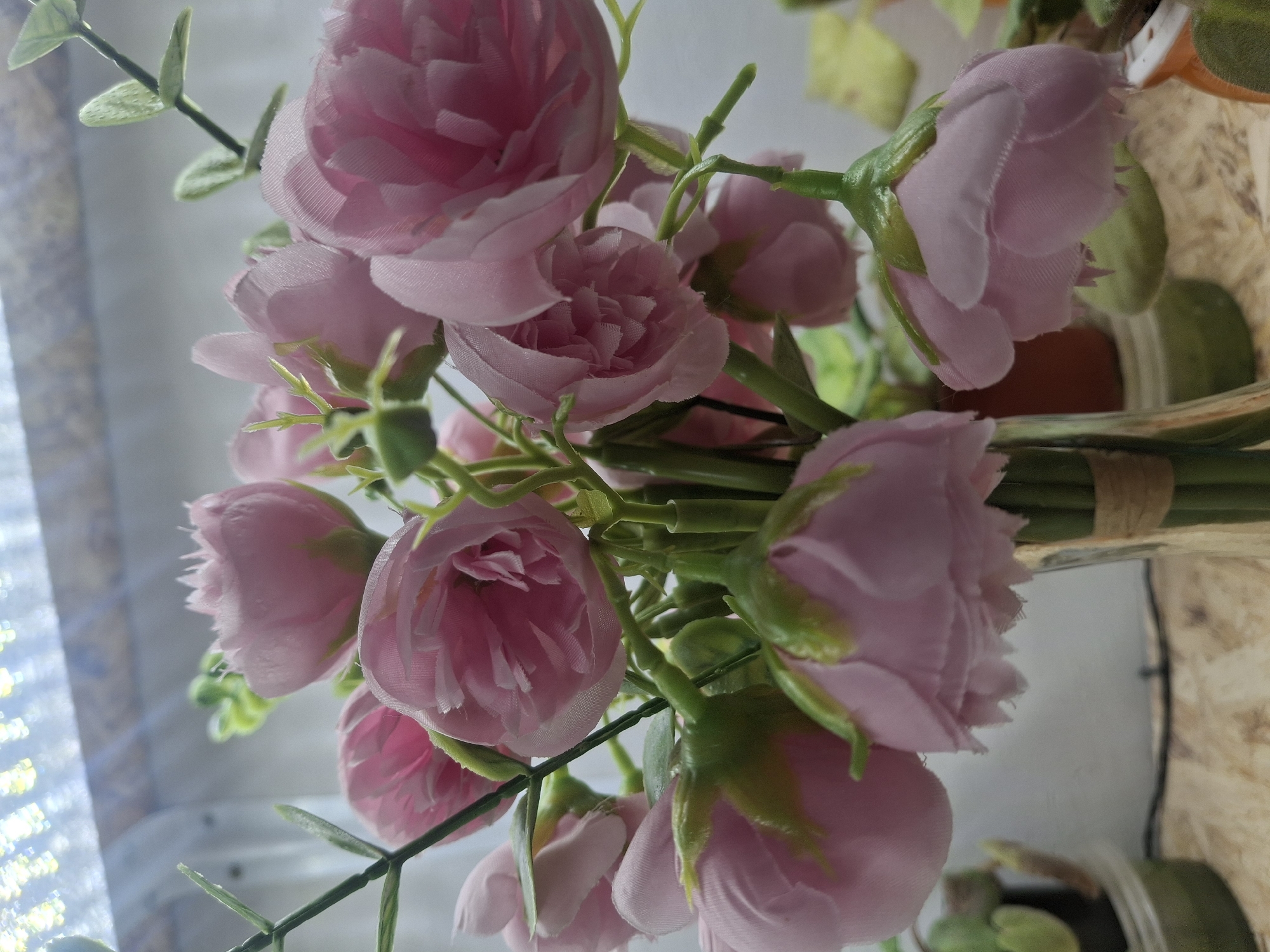 The image shows a close-up of a bouquet of artificial pink roses in a glass vase. The roses have multiple layers of soft pink petals and green stems with small leaves. The vase is tied with a piece of twine. In the background, there are out-of-focus plants in pots, and a bright light source that appears to be an LED grow light. The surface the vase is resting on appears to be a light-colored wood grain. There are no people or animals visible in the image, and no specific geographical features are identifiable that would place the scene in Suzak, Kyrgyzstan. The focus is on the floral arrangement and the lighting.