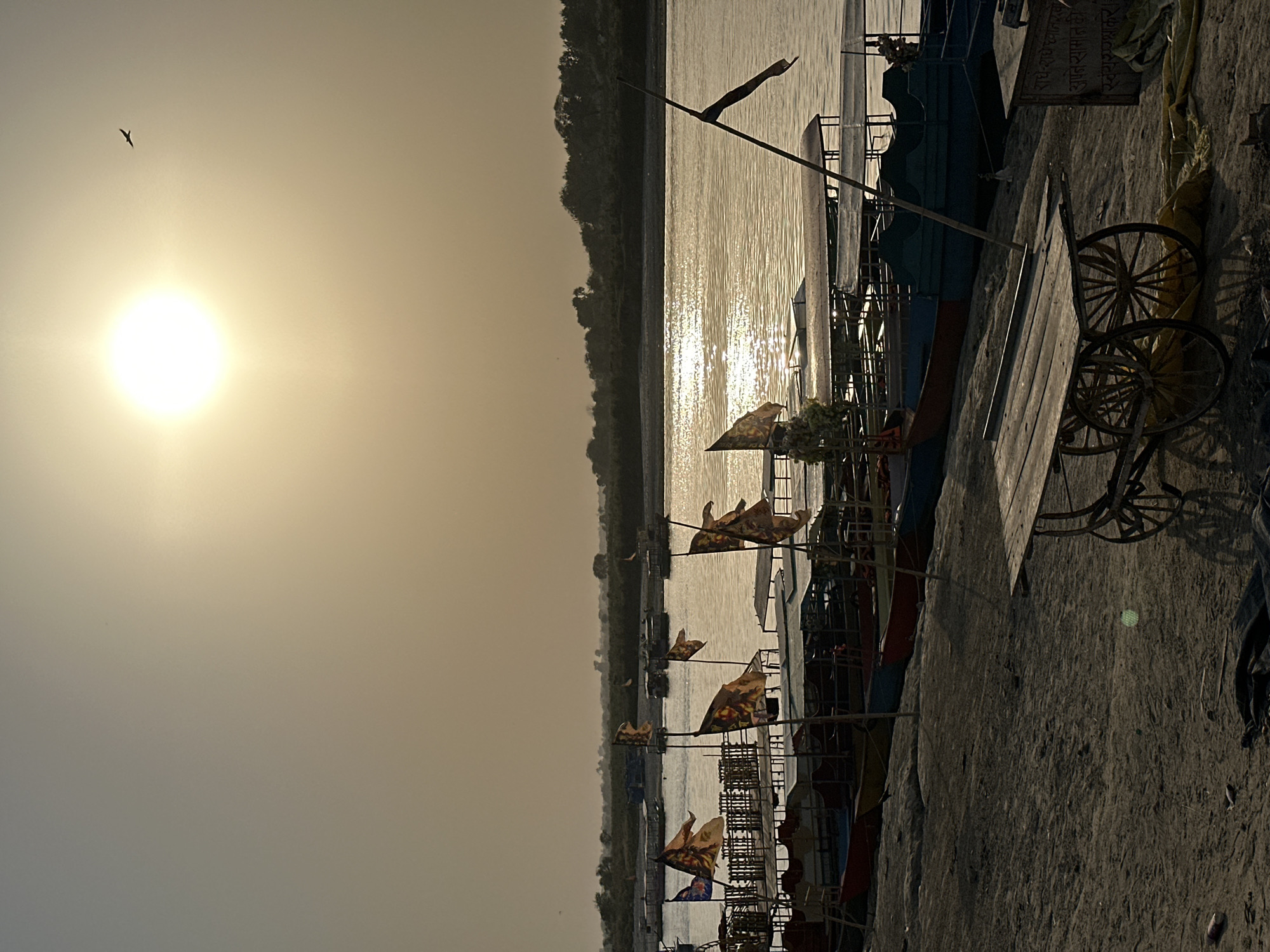 This is an outdoor, daytime shot taken near the Yamuna River in Vrindavan, India. The low sun reflects off the water's surface. On the right side of the frame, there are several small boats docked or tied to the riverbank. These boats are adorned with flags and religious symbols. Adjacent to the boats, there is a wooden cart with spoked wheels. The foreground shows the ground or riverbank, which appears to be composed of dirt or sand. In the background, across the river, a densely vegetated area lines the opposite bank. High in the sky, a bird is seen flying.