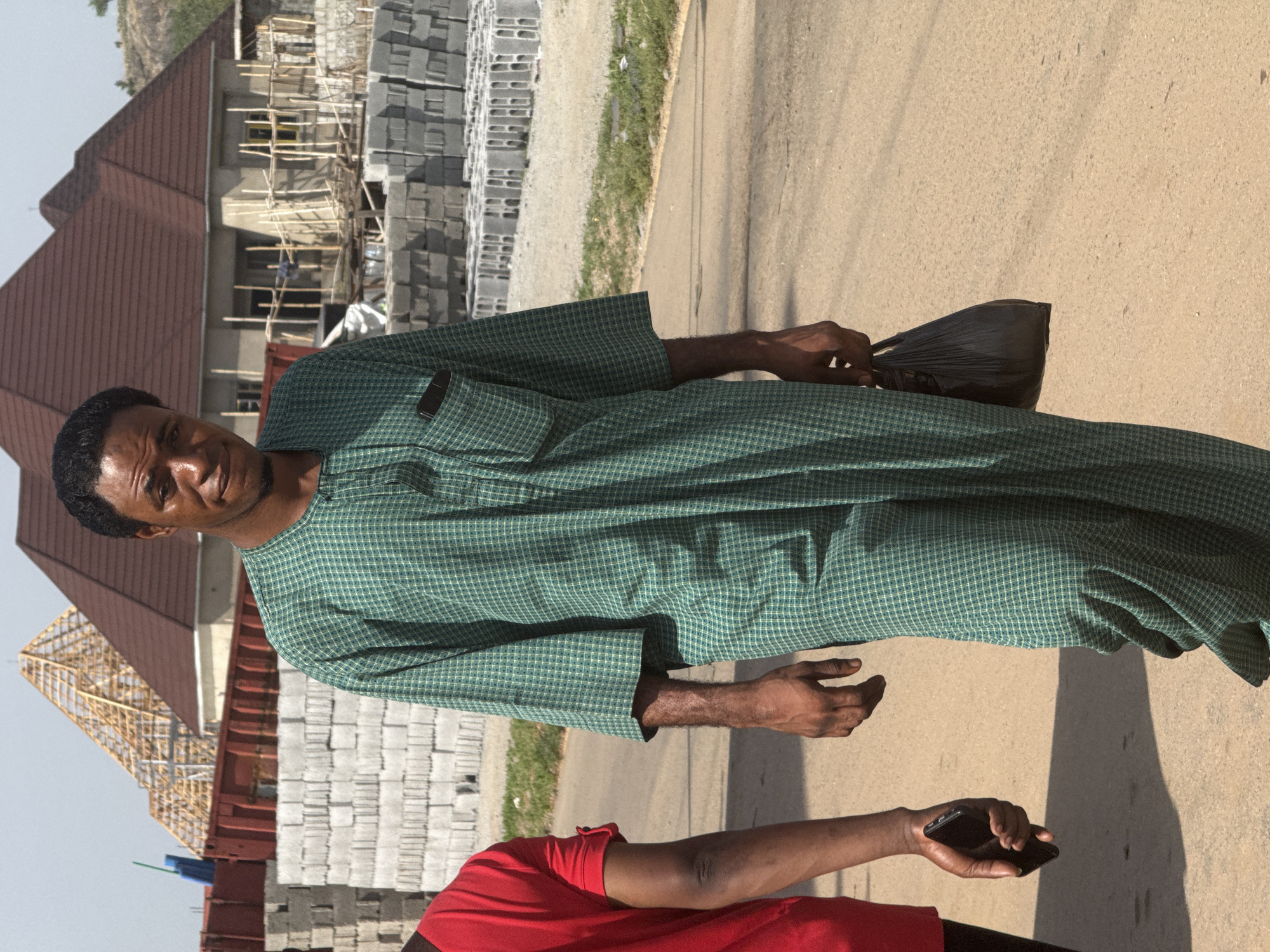The image captures an outdoor scene in Abuja, Nigeria, featuring a man in the foreground. He is dark-skinned, with short dark hair, a light mustache, and a small beard. He wears a traditional-style long-sleeved tunic, possibly an Agbada, in a green and white checkered pattern. He is looking directly at the camera with a faint, friendly smile, and holds a black plastic bag in his right hand.
Partially visible in the lower right of the frame is another person, wearing a red t-shirt. Their arm and hand are visible, holding a black smartphone.
The background shows a dynamic urban landscape. To the man's left, there are several buildings, some with red-brown sloped roofs, suggesting residential structures. Behind him and to his right, there's an active construction site with a building under scaffolding and numerous grey concrete blocks neatly stacked on the ground. Beyond the buildings, a distant rocky hill or mountain range is visible under a clear sky.
The ground is a light-colored paved surface, likely a road or a wide pathway, with distinct shadows cast by the subjects and unseen objects, indicating bright sunlight and a clear day, likely midday or afternoon. The overall impression is a bustling area undergoing development, characteristic of parts of Abuja. No visible text is present in the image.