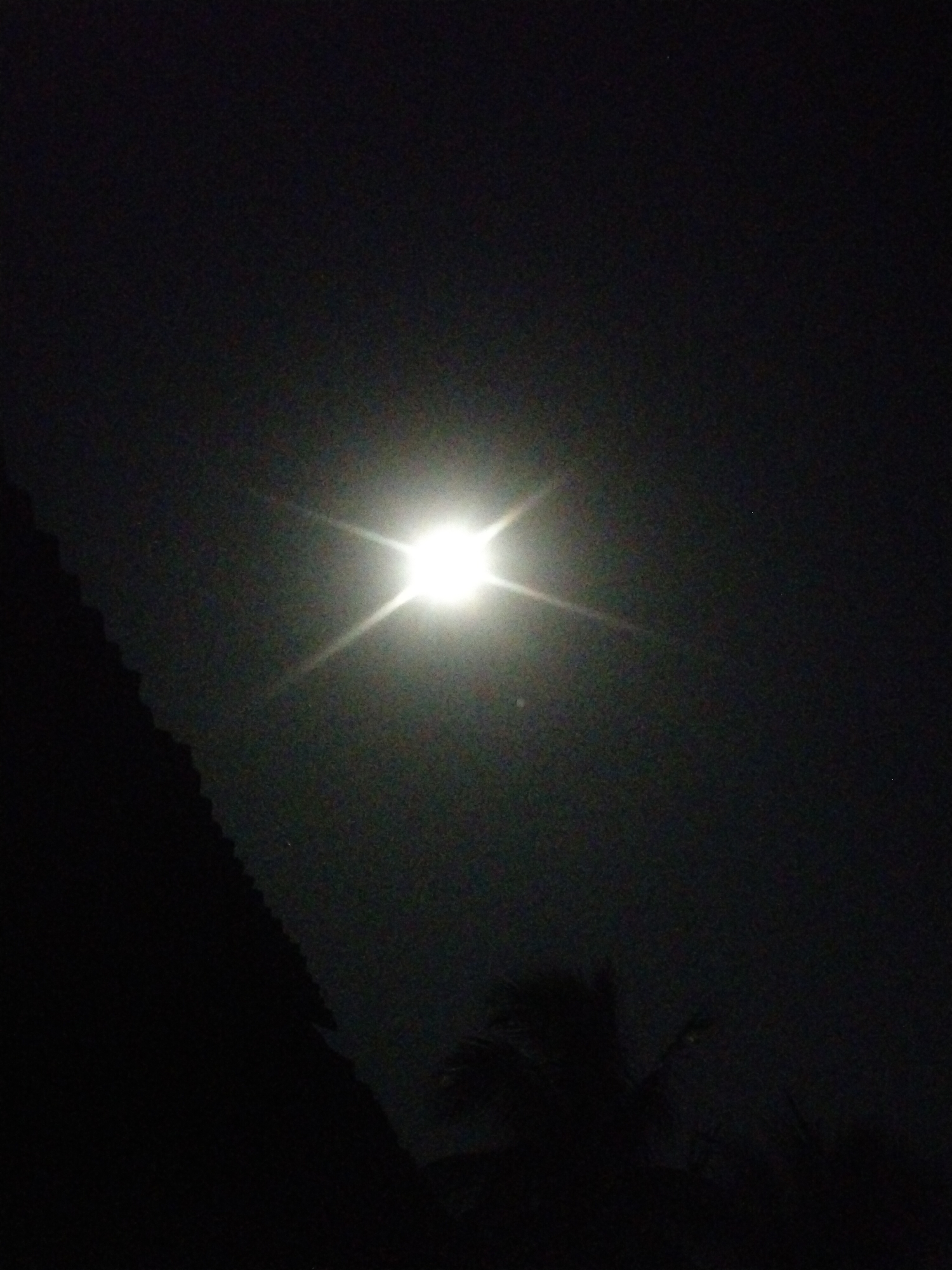 The image shows a night scene in Uyo, Nigeria, with a bright light source, likely the moon, dominating the sky. The lower portion of the frame contains dark silhouettes of trees and possibly a building. The sky is dark with some visible texture, suggesting either a cloud cover or atmospheric particles. The scene appears to be largely devoid of human activity, focusing on the natural elements of light and foliage in the darkness.