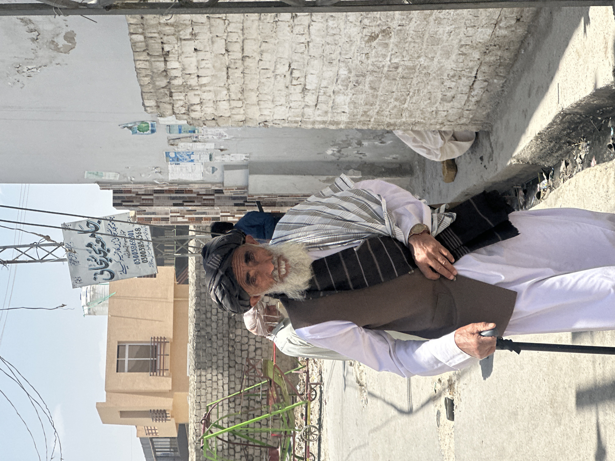 The image captures an elderly man smiling brightly on a sunlit street in Quetta City Tehsil, Pakistan. He is the central figure, dressed in traditional white clothing (shalwar kameez), a dark vest, and a dark turban, with a prominent white beard. He holds a black walking stick.
Behind him, another person is partially visible, and further down the street, a third individual can be seen. The street appears to be an urban or semi-urban alleyway, with various buildings lining both sides, some with exposed brick and others plastered. Overhead wires are visible against the clear sky. Strong shadows suggest bright midday sun. On the ground, there is some debris and a small, colorful metal structure, possibly a playground item or part of a cart.
A prominent sign hangs overhead, featuring text in Urdu/Arabic script. The text reads: "In the name of Allah, the Most Gracious, the Most Merciful, O Ever-Living, O Self-Subsisting. Jamia Masjid Sajjani. For good deeds in Jamia Masjid Sajjani. Trustee Syed Haji Wali Shah. Contact numbers: 03003869601, 03003854368. Located near Maid Colony, Central Road, Wata." This sign likely serves as an announcement or an appeal for charity related to a mosque.