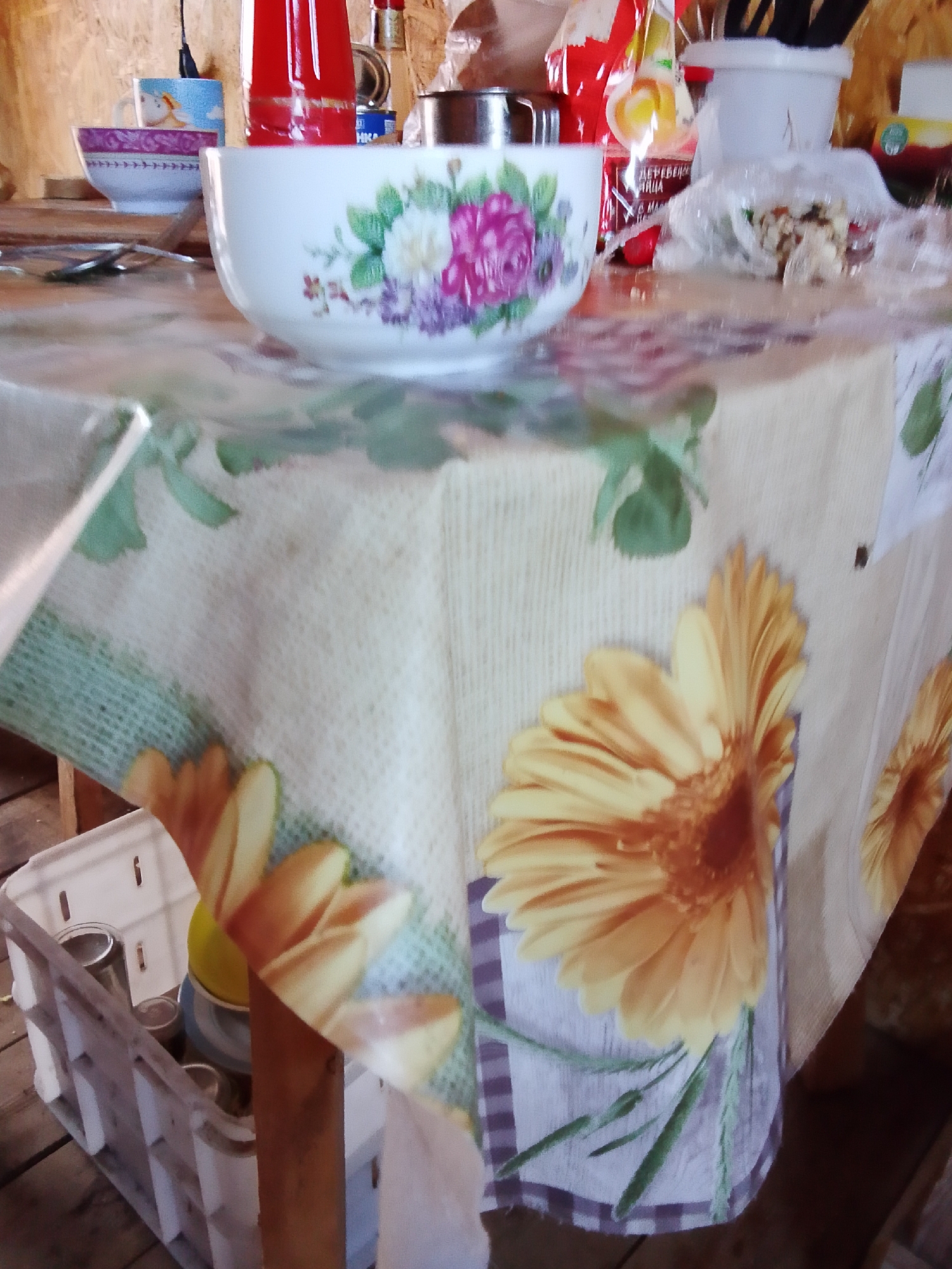The image depicts a close-up view of a table covered with a tablecloth featuring a floral and checkered pattern. On the table, there is a white bowl decorated with colorful flowers, and to its left, a smaller bowl with a purple rim and a pattern of what appears to be a cartoon character. Further back, various items are visible, including bottles, a metal pot, and plastic packaging. To the left of the table, a white plastic crate contains several glass jars and a yellow object. The overall setting appears to be rustic, with wooden elements visible in the background and on the floor. The scene does not provide any specific information about the city of Yelanka, Russia.