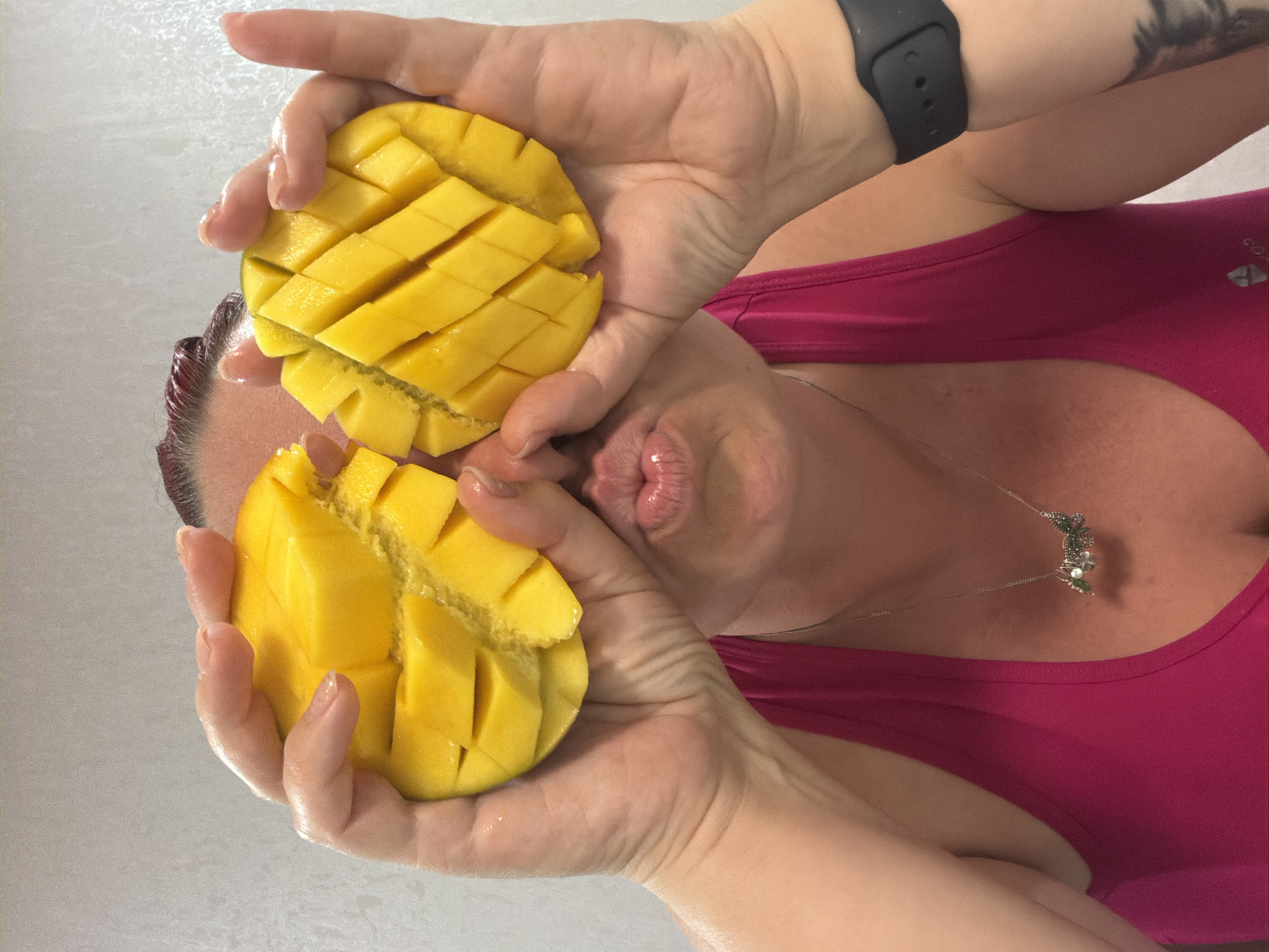 A woman in a pink tank top holds two halves of a mango in front of her face. The mangoes are cut into a grid pattern, exposing the yellow flesh. The woman's lips are pursed. She is wearing a necklace with a floral pendant and a black smartwatch on her wrist. Her hair is visible at the top left, with a reddish tint. The background is a plain, light-colored wall. There is no indication of the city of Chaikovsky, Russia.