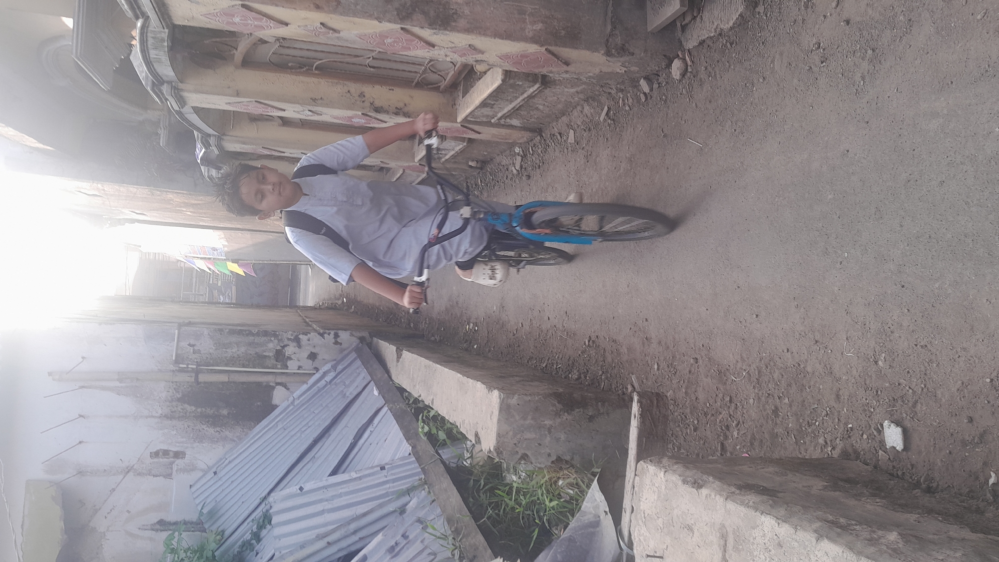 A young boy rides a bicycle down a narrow dirt path between two weathered buildings. He is wearing a light blue traditional outfit and a backpack. The buildings have peeling paint and some exposed brickwork. The path is lined with a low concrete wall on the left, where some corrugated metal sheeting is piled. On the right, the wall of a building displays a decorative pattern. The scene is lit by bright sunlight, which creates some glare in the upper portion of the image. The image was taken in Tasikmalaya, Indonesia.