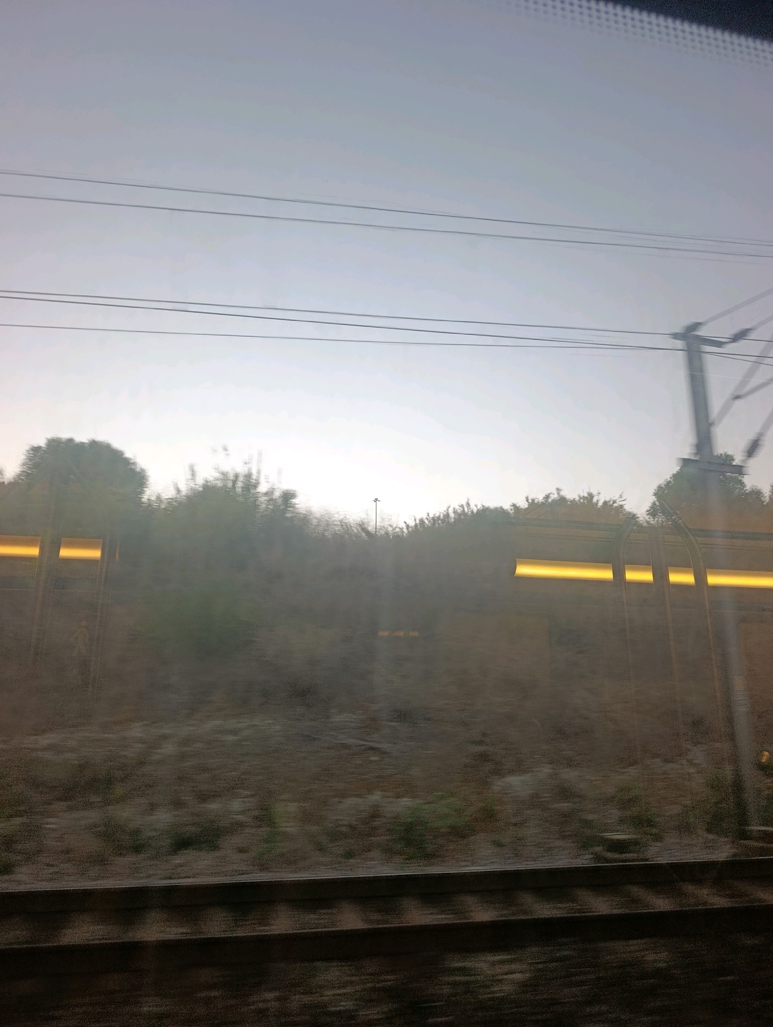 The image is a blurred, outdoor scene taken from a moving vehicle, likely a train, looking out a window. The time of day appears to be either early morning or late evening due to the low light and hazy sky. The foreground shows what appear to be train tracks and the embankment beside them. In the middle ground, there is a slope covered in dry vegetation and some trees, with horizontal yellow lights visible at intervals, possibly from the side of the train or infrastructure. In the background, the sky is a pale, washed-out gray, with visible overhead power lines. A blurred silhouette of a utility pole with crossbars is visible on the right side. There are no people or discernible actions visible in the image. The scene does not contain any identifiable landmarks or features that would indicate a specific location within the city of Almada, Portugal.