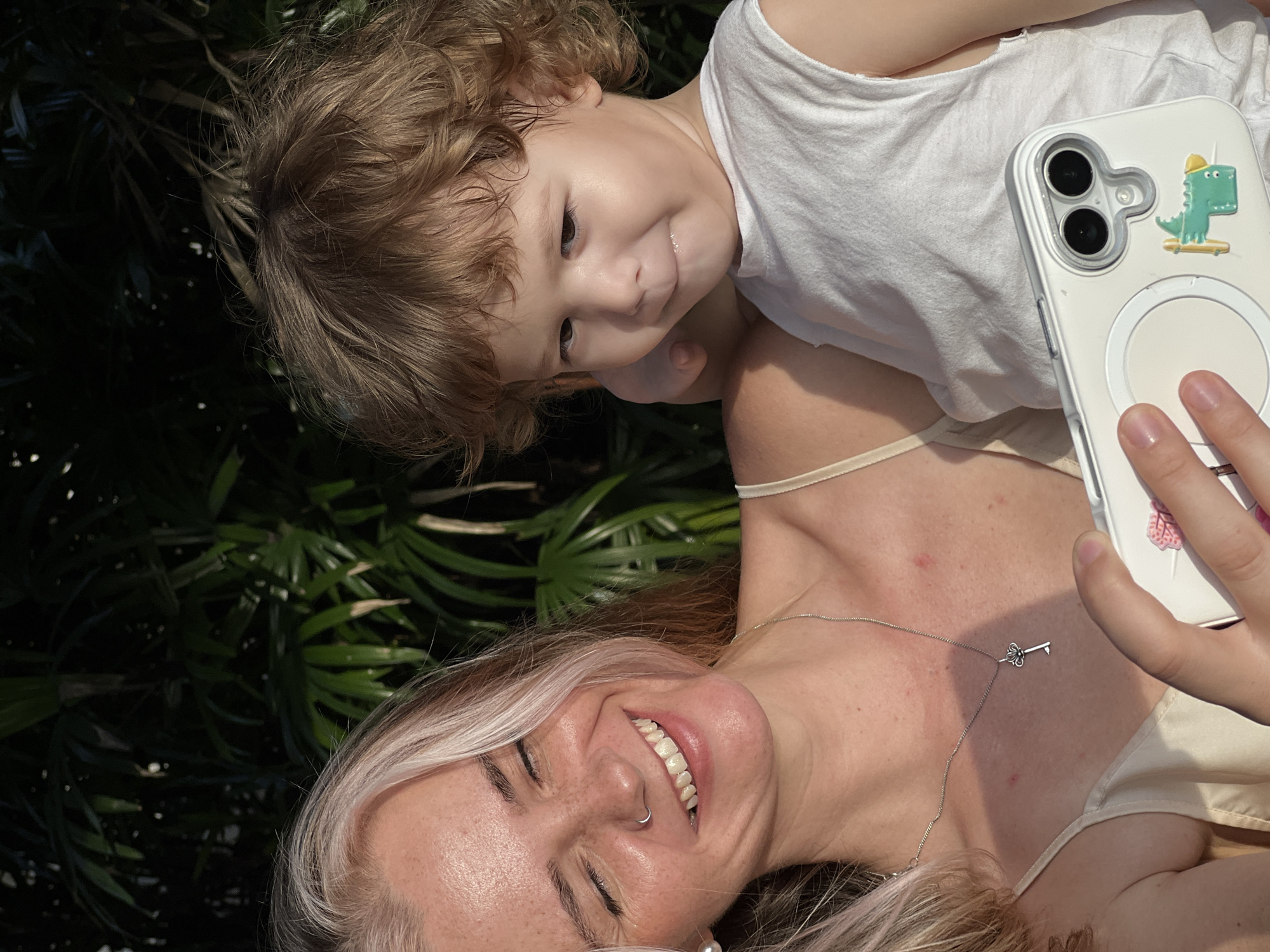 A medium shot captures a woman and a child outdoors, with lush green foliage in the background. The woman, positioned on the left, is smiling with her eyes closed and looking down at a smartphone she is holding. She has blonde hair with streaks of lighter blonde, freckles, and wears a cream-colored tank top and a delicate silver necklace with a key pendant. The child, on the right, is looking slightly to their left with a playful expression, their mouth slightly open, and wears a white sleeveless shirt. The child is held close by the woman, with their left arm resting on the woman's shoulder. The smartphone in the woman's hand has a white case with a circular magnetic accessory and a cartoon dinosaur sticker. The lighting suggests it is either early morning or late afternoon, casting a warm glow on the subjects.