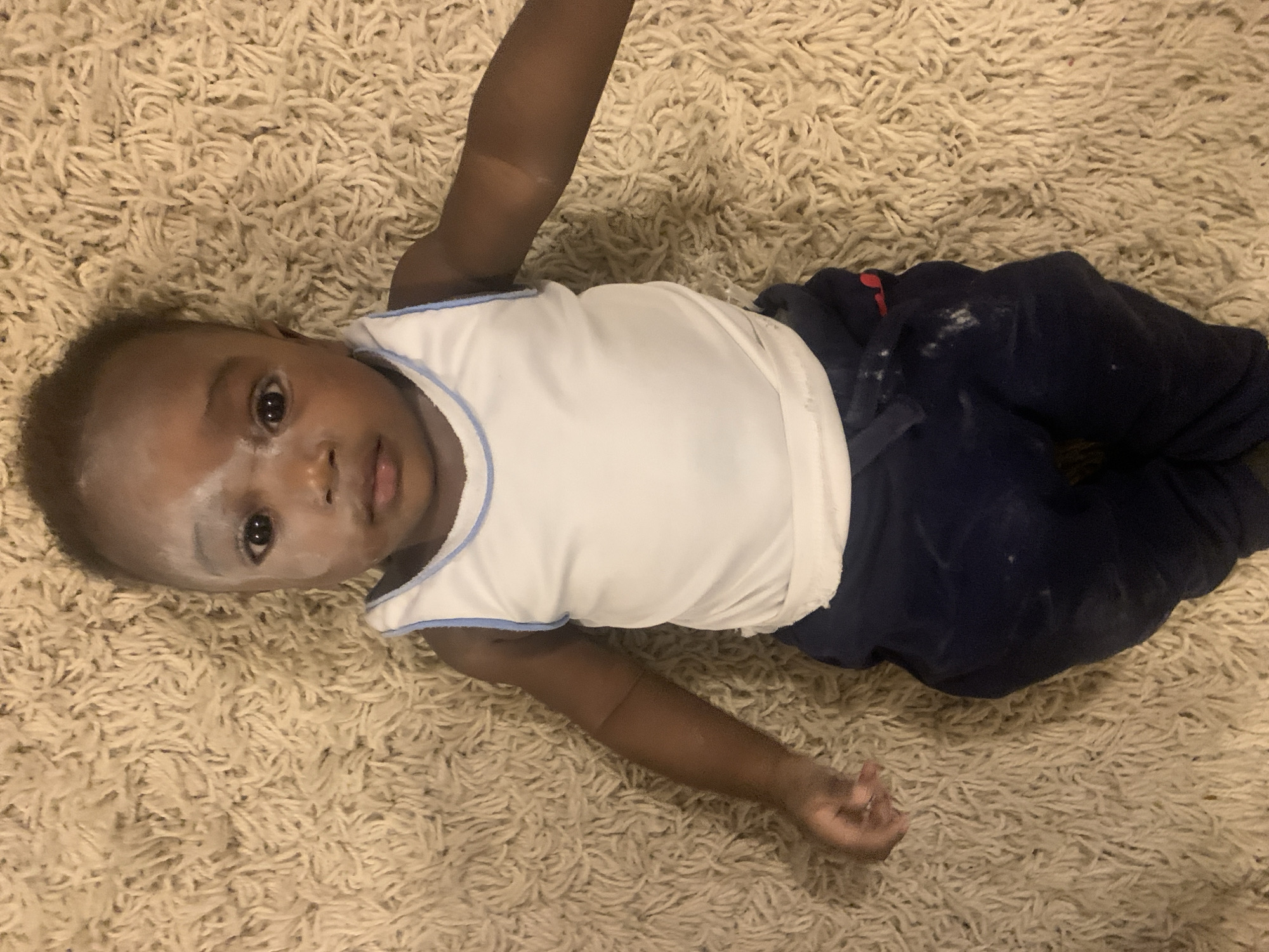 A dark-skinned baby lies on their back on a beige shag carpet. The baby wears a white tank top with blue trim and dark blue sweatpants. A white powdery substance is smeared across the baby's face. The location is unknown and there is no evidence to suggest it is in Barakin Akawo, Nigeria.