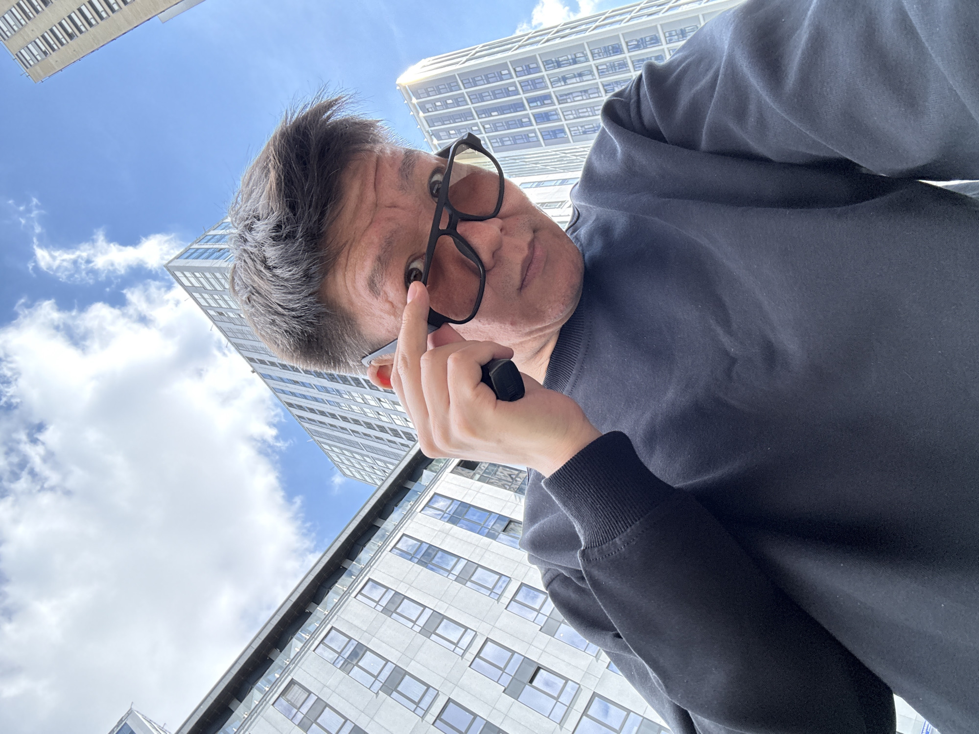 An upward-facing selfie is taken by a person outdoors, with modern high-rise buildings and a blue sky with clouds in the background. The person, appearing to be of East Asian descent, is wearing a black crewneck sweatshirt and dark-rimmed sunglasses with tinted lenses. They are looking directly at the camera and adjusting their sunglasses with their right hand. The architecture suggests an urban environment, consistent with a location in Moscow, Russia.