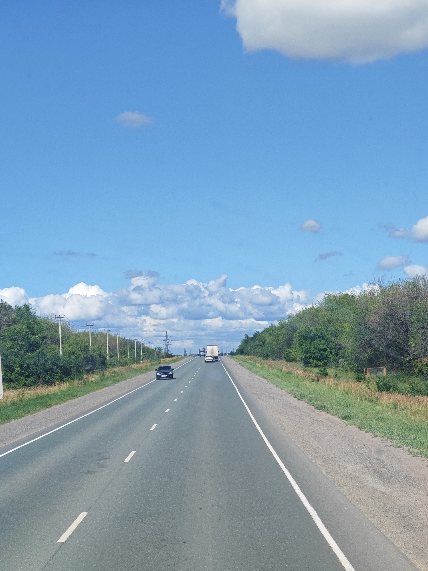 The image captures a view from a vehicle traveling on a paved highway under a clear blue sky with scattered white clouds. The road is a two-lane highway, with a white solid line on the left and dashed white lines dividing the lanes. The highway is flanked by green trees and vegetation on both sides. There are utility poles visible along the left side of the road. In the distance, several vehicles, including cars and trucks, are visible on the highway. The scene suggests a rural or semi-rural setting, possibly in Maloe Tomylovo, Russia, given the context.